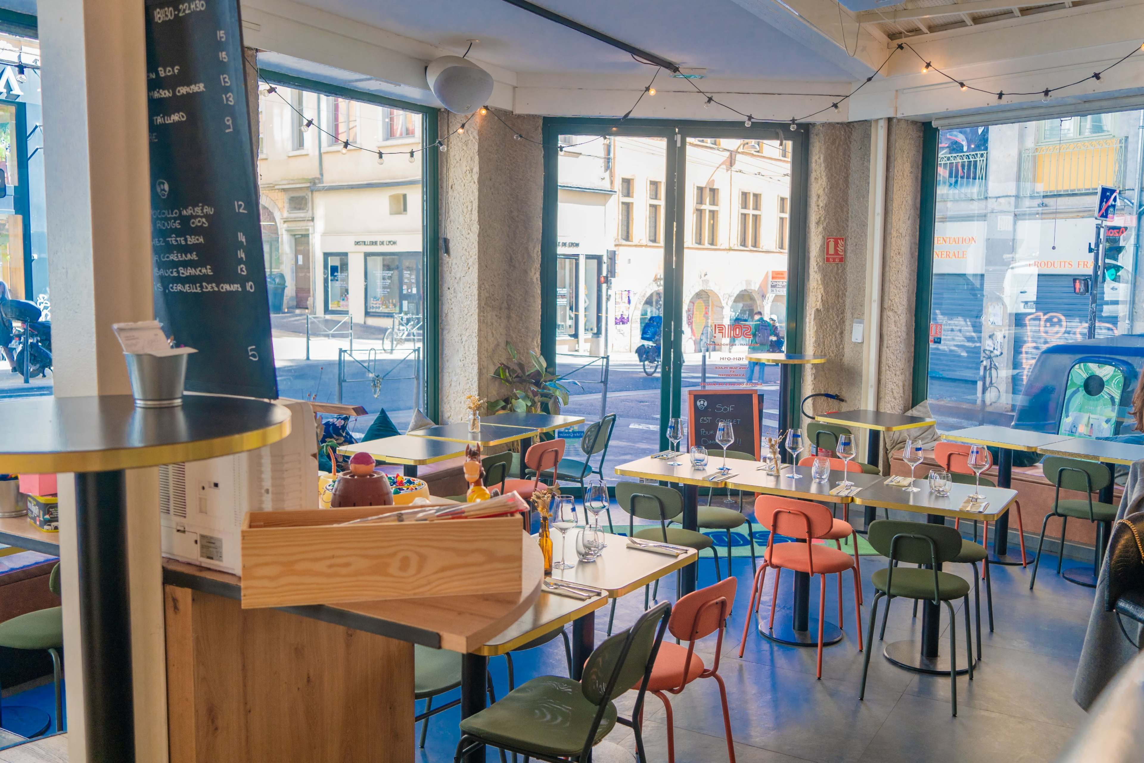 The image shows a modern café interior with colorful chairs and tables, large windows displaying a street view, and a menu board on a central pillar.