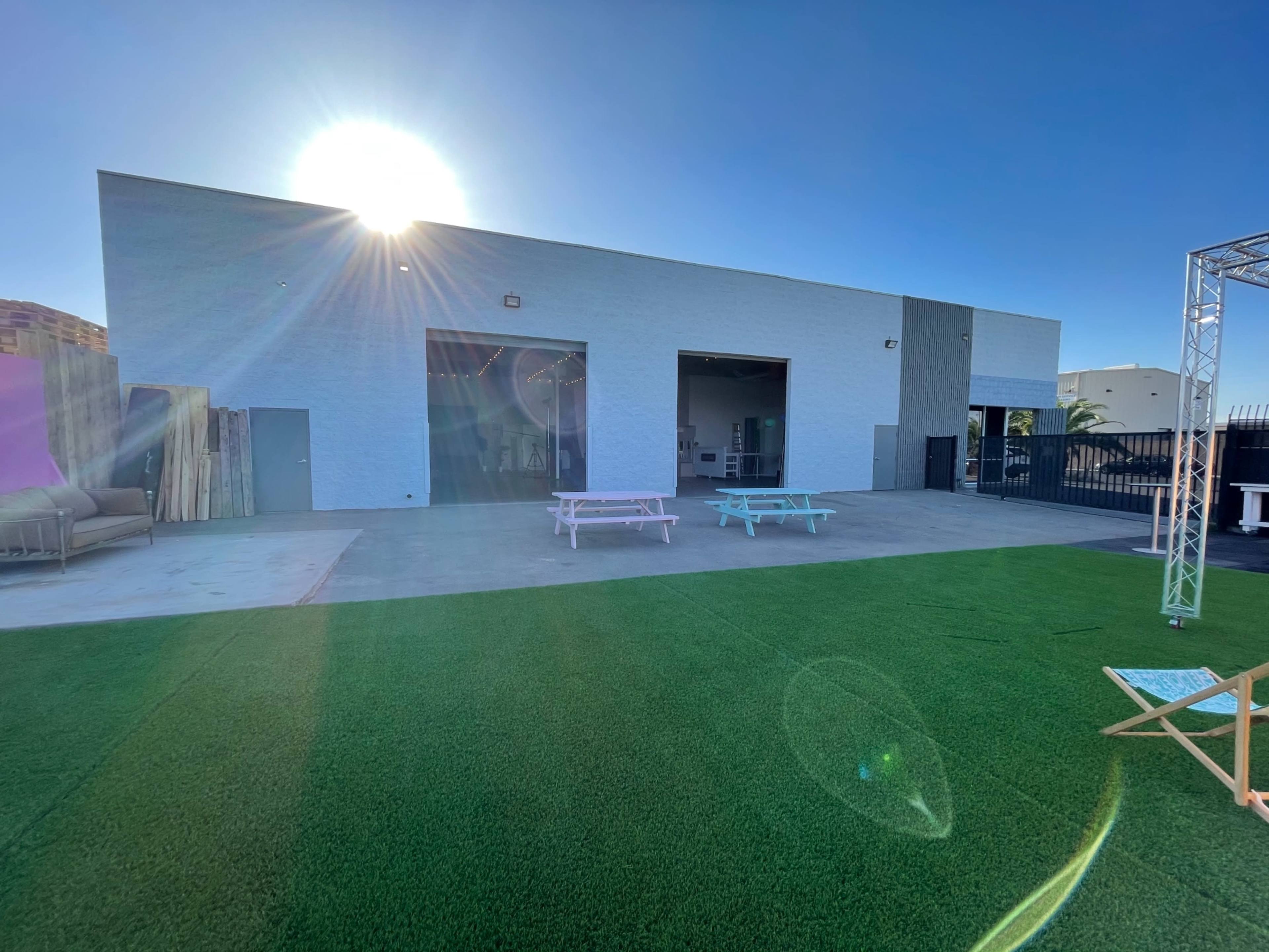 A light-colored building with large doors is set against a clear blue sky, with green artificial grass and patio furniture in the foreground.