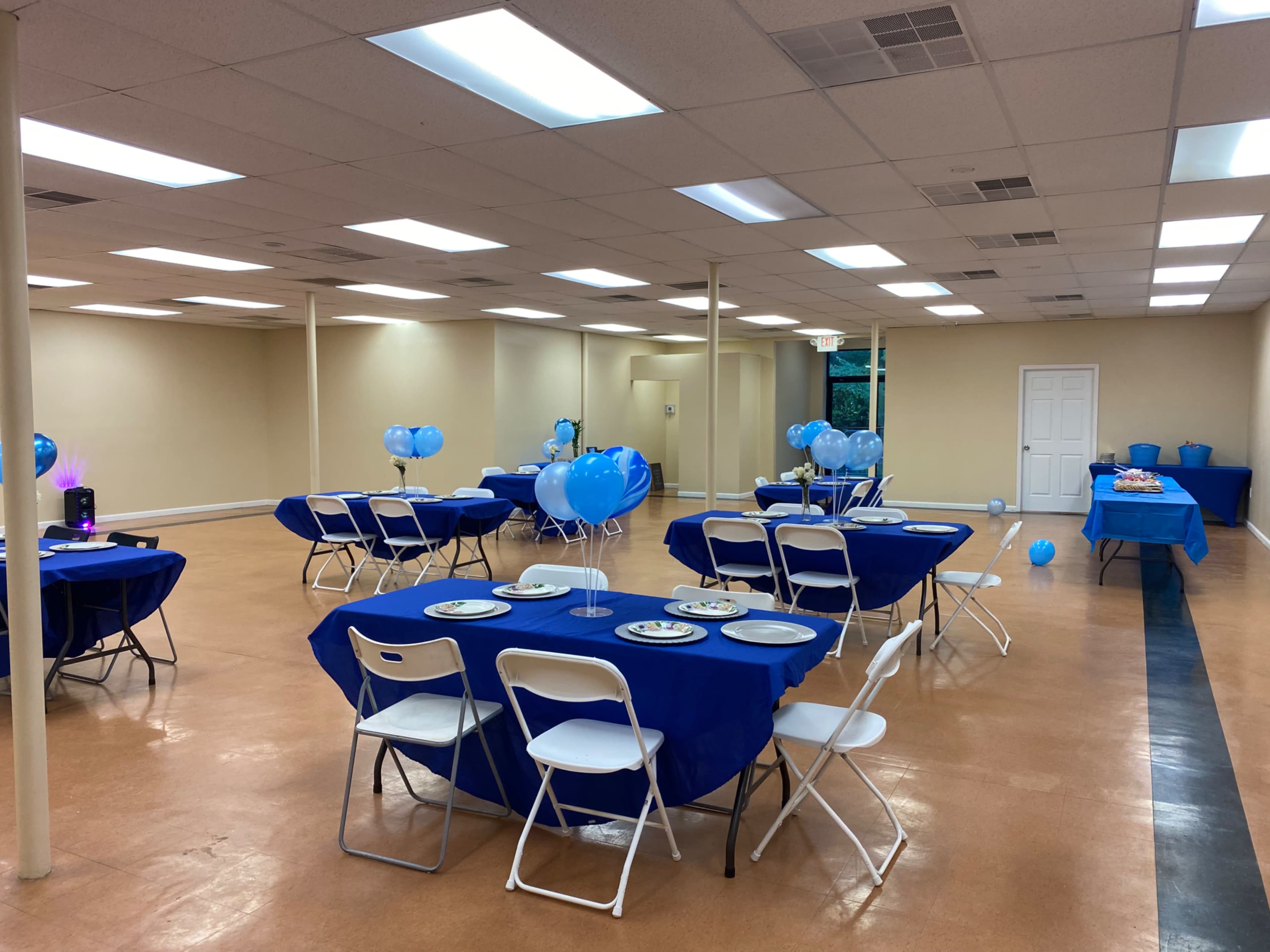 The image shows a spacious room arranged with tables covered in blue tablecloths, white chairs, and blue balloons scattered around.