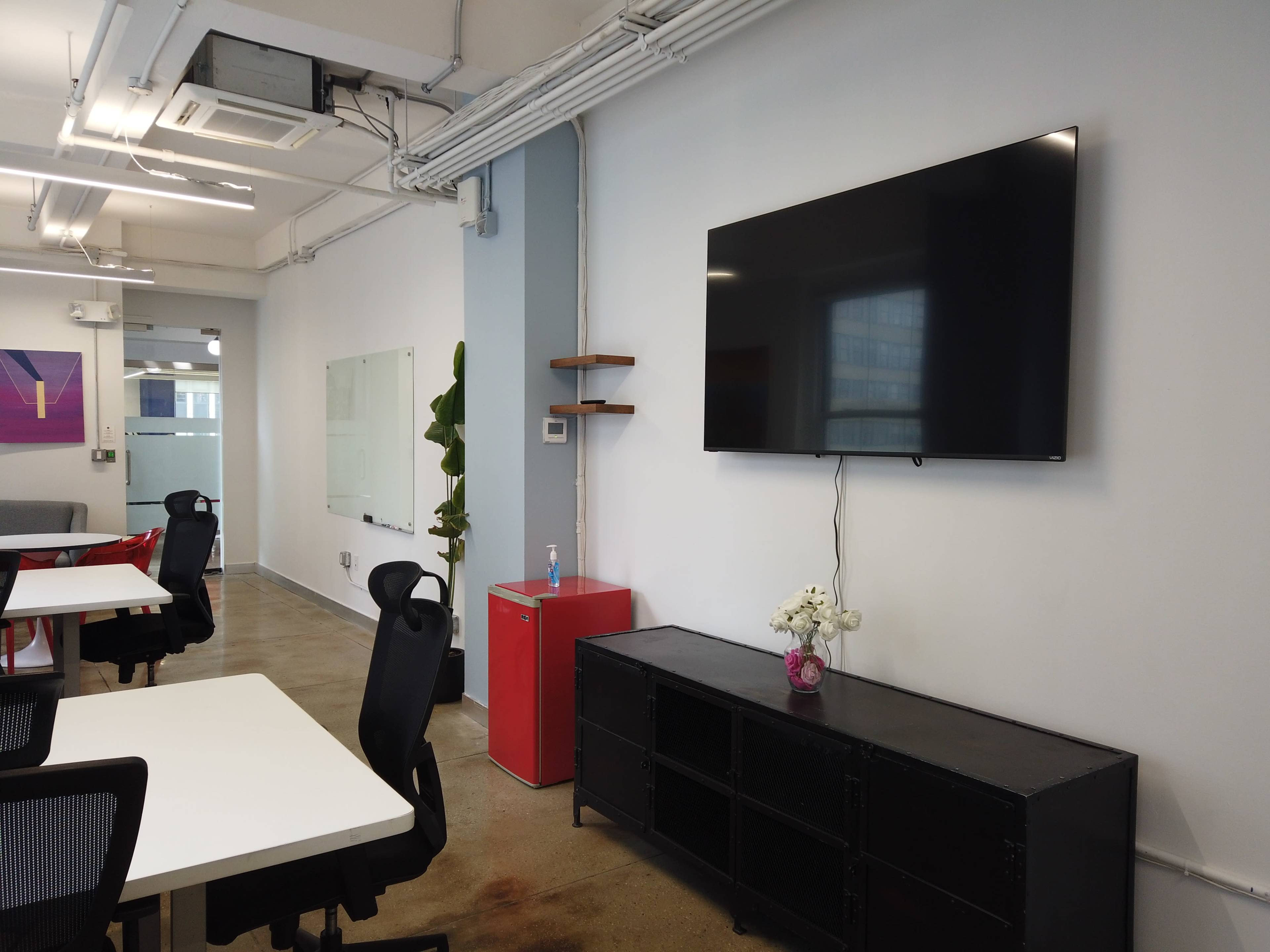 A modern office space features a large wall-mounted television, a black storage unit, and a small red fridge, with white tables and black chairs arranged for work.