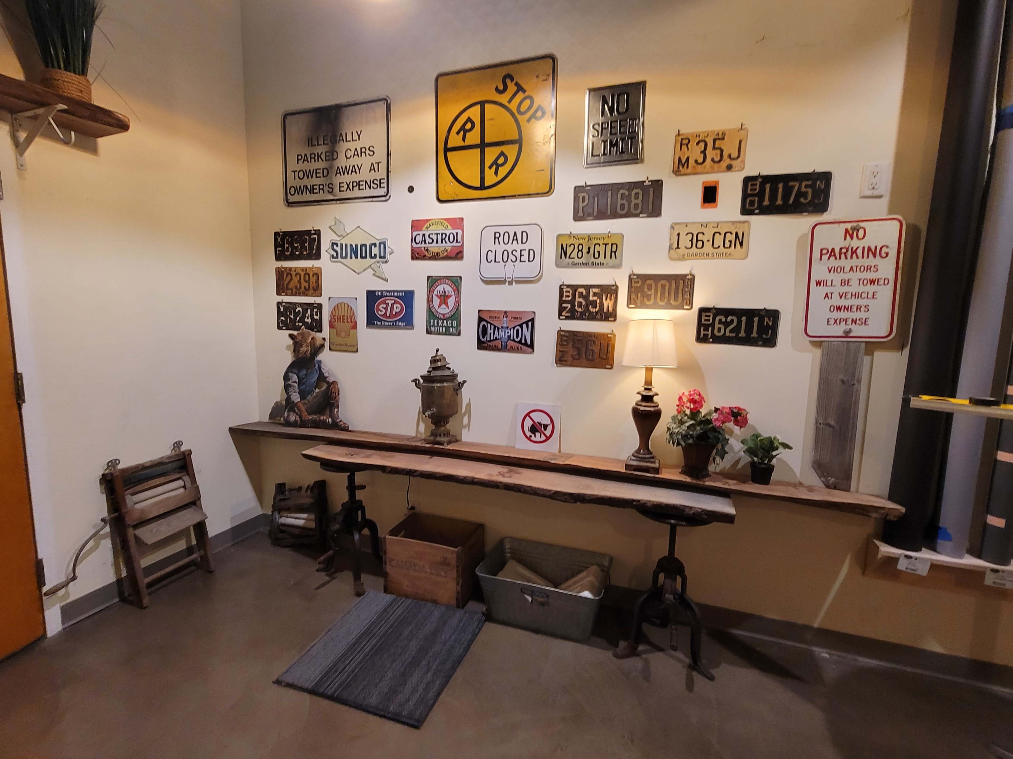 The image shows a corner of a room featuring a collection of vintage signs on a wall, a wooden bench, a lamp, and decorative flowers.