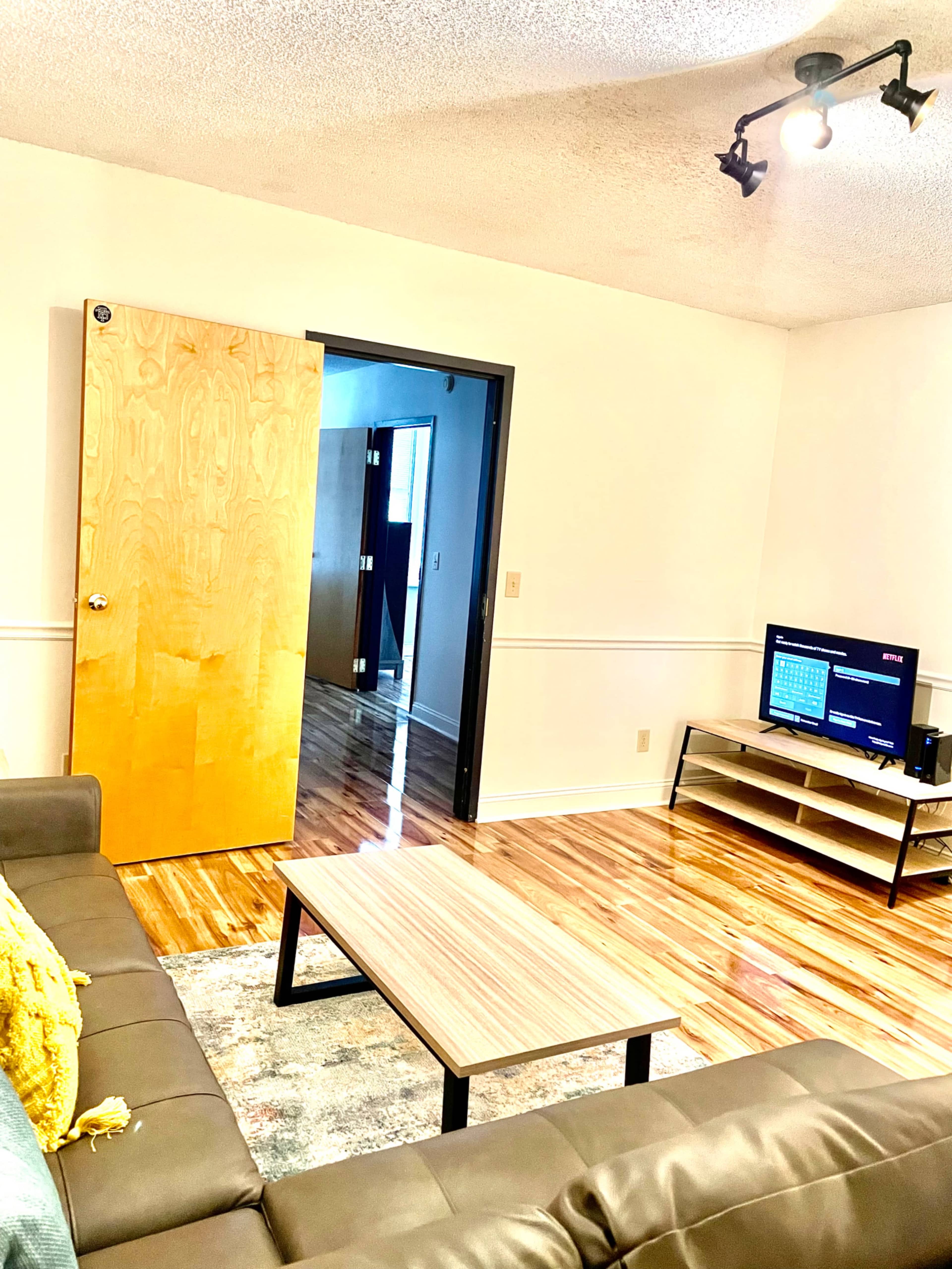 The living room features a brown couch, a wooden coffee table, and a television on a stand, with a door leading to a hallway in the background.