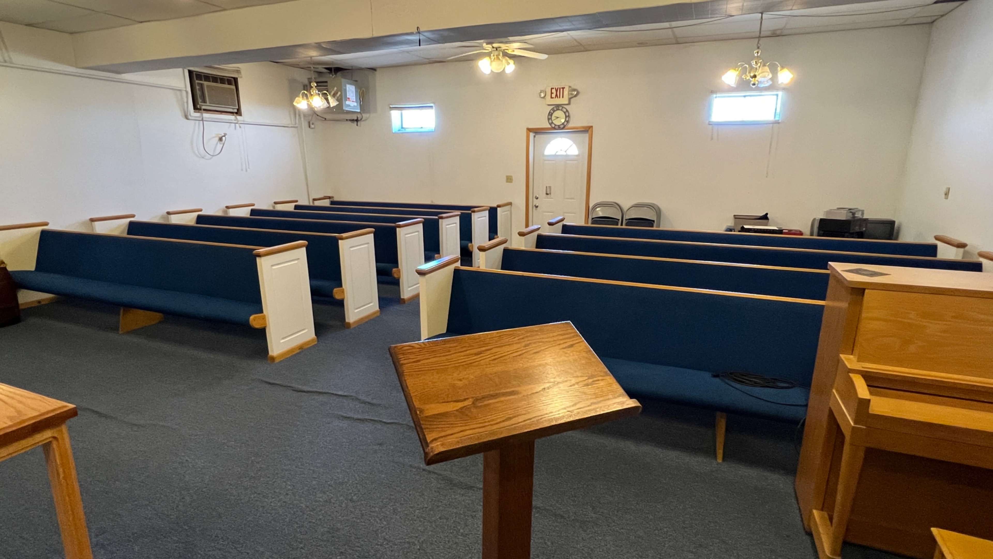 The interior of a small church or meeting space with wooden pews arranged in rows and a podium at the front.