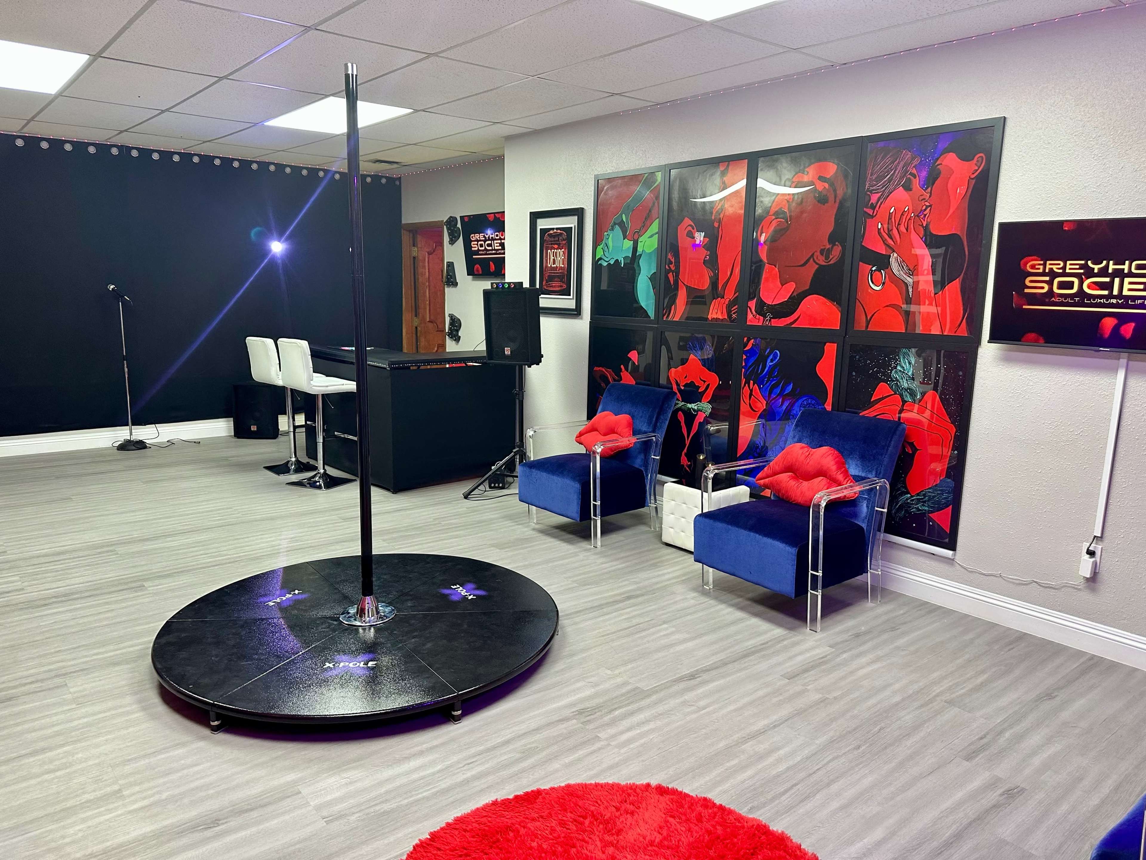 A modern studio space featuring a dance pole on a circular platform, two blue chairs with red cushions, and a vibrant wall adorned with framed artwork.