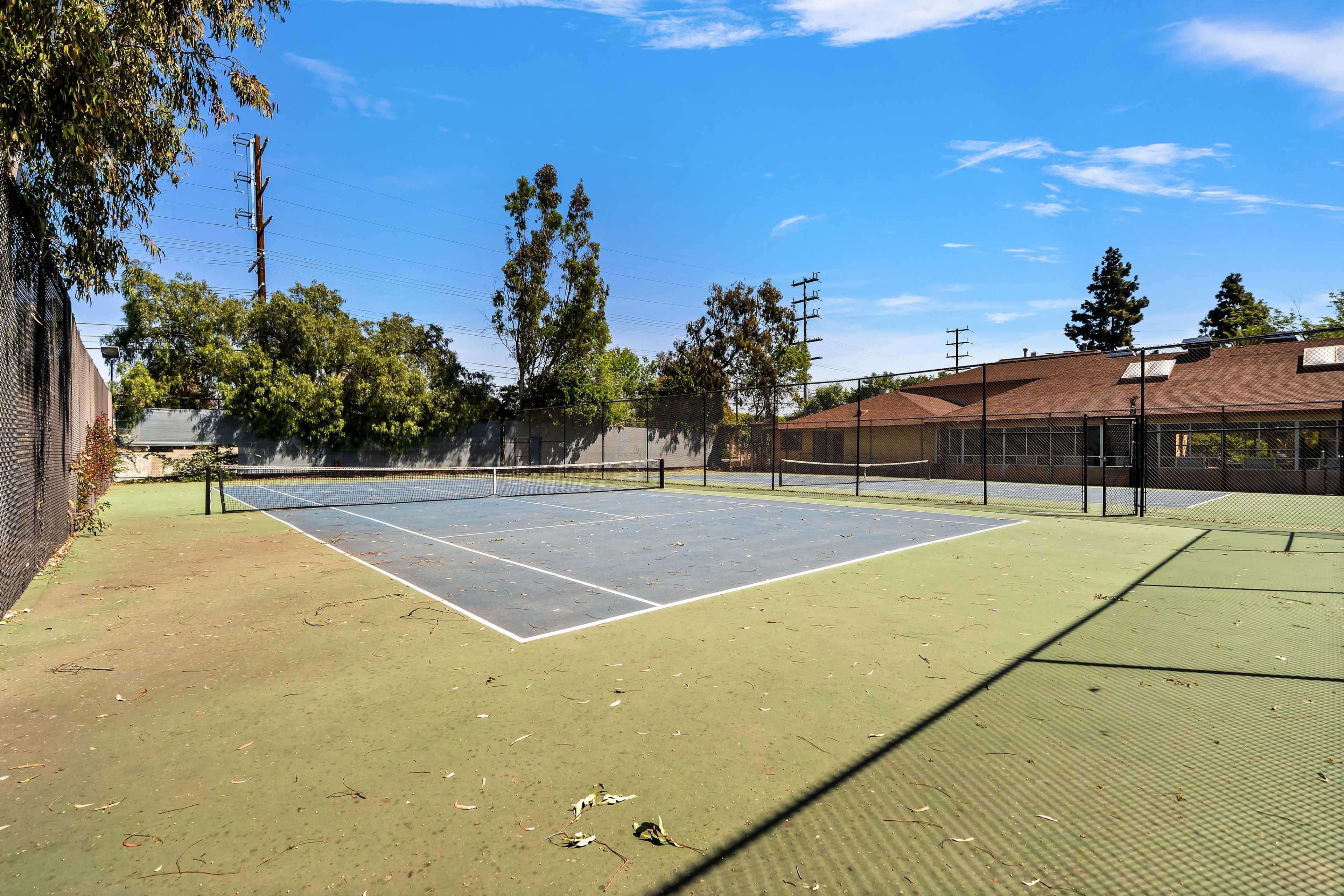 An empty, outdoor tennis court with a blue and green surface surrounded by trees and a fence.