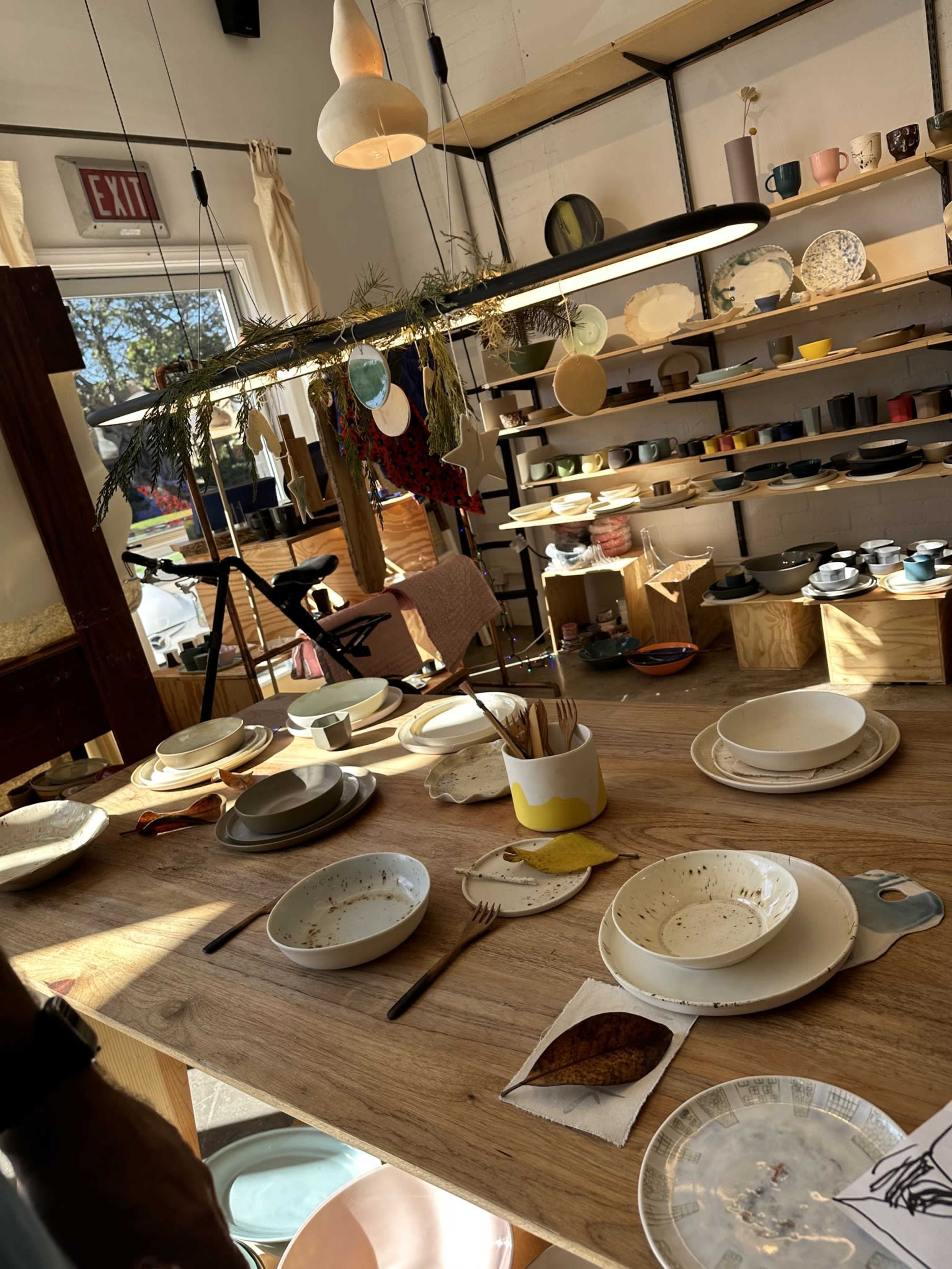 The image shows a brightly lit table cluttered with used dishes, utensils, and decorative items, surrounded by shelves displaying an assortment of colorful ceramic cups and plates.