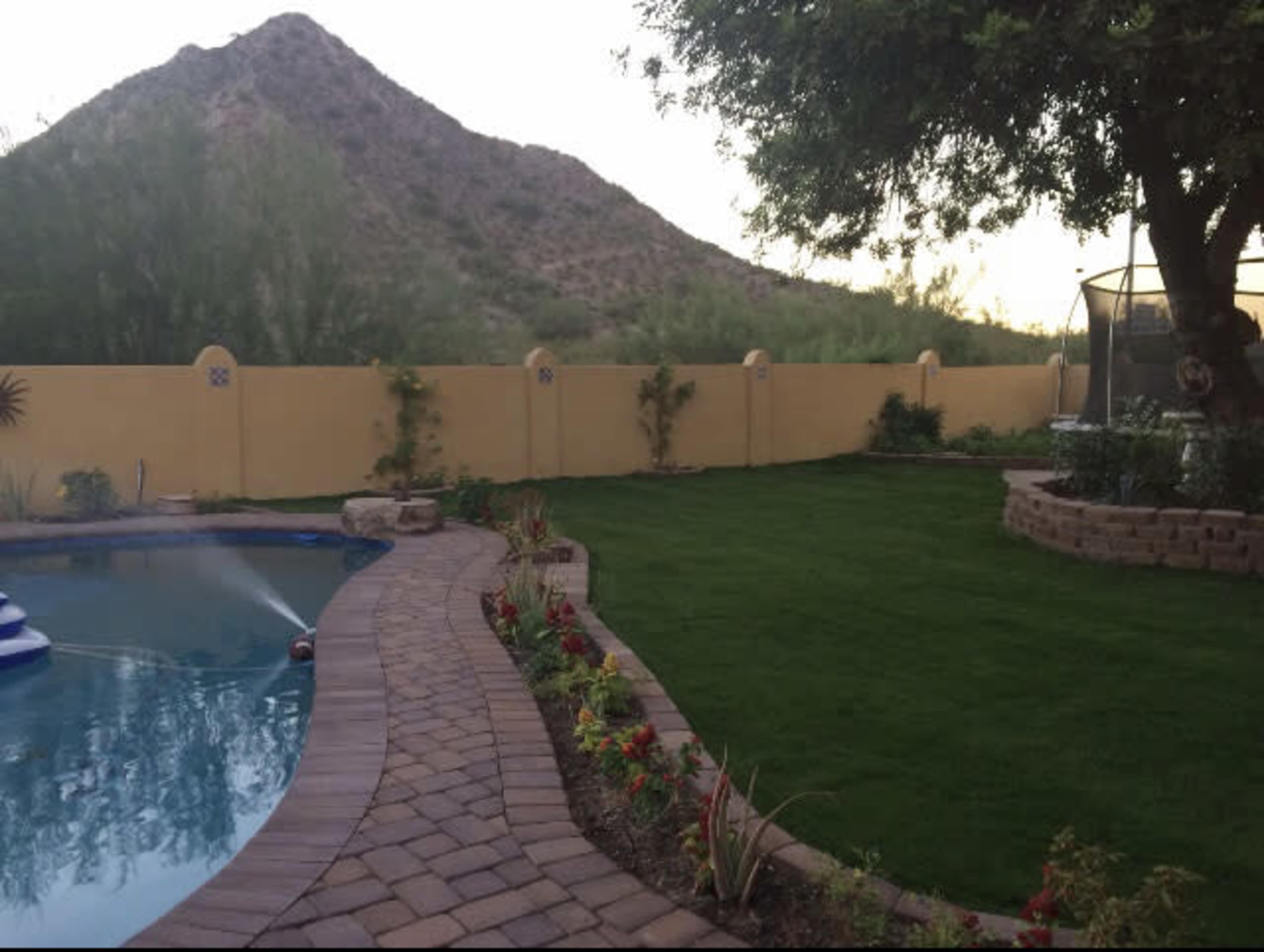 The image shows a landscaped backyard with a swimming pool, a stone pathway, lush green grass, and a mountain in the background.