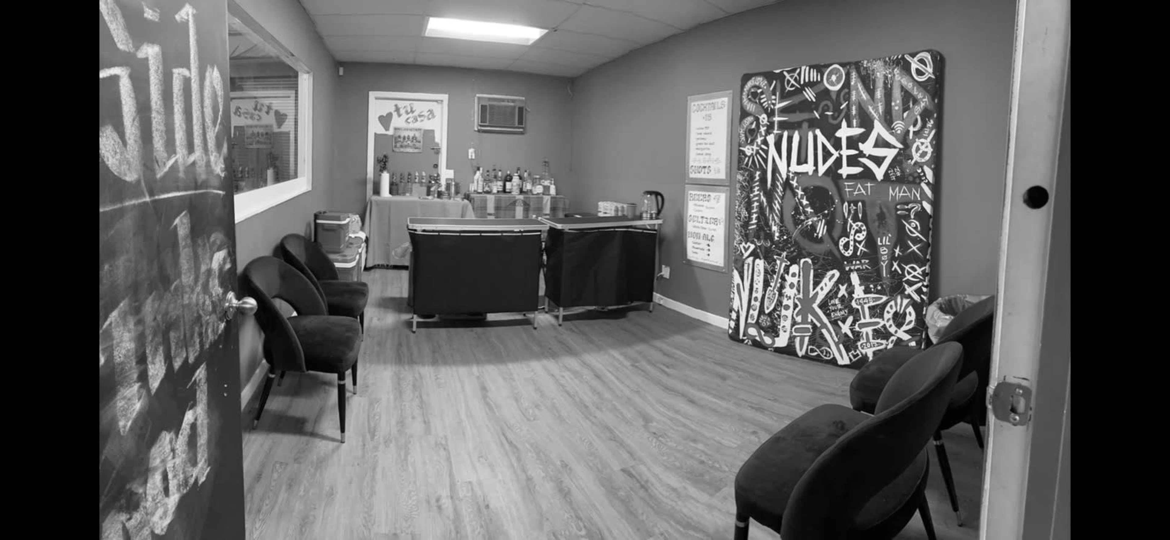 The image shows a room with wooden flooring, a bar counter with bottles behind it, several black chairs arranged in the foreground, and a large mural on one wall.
