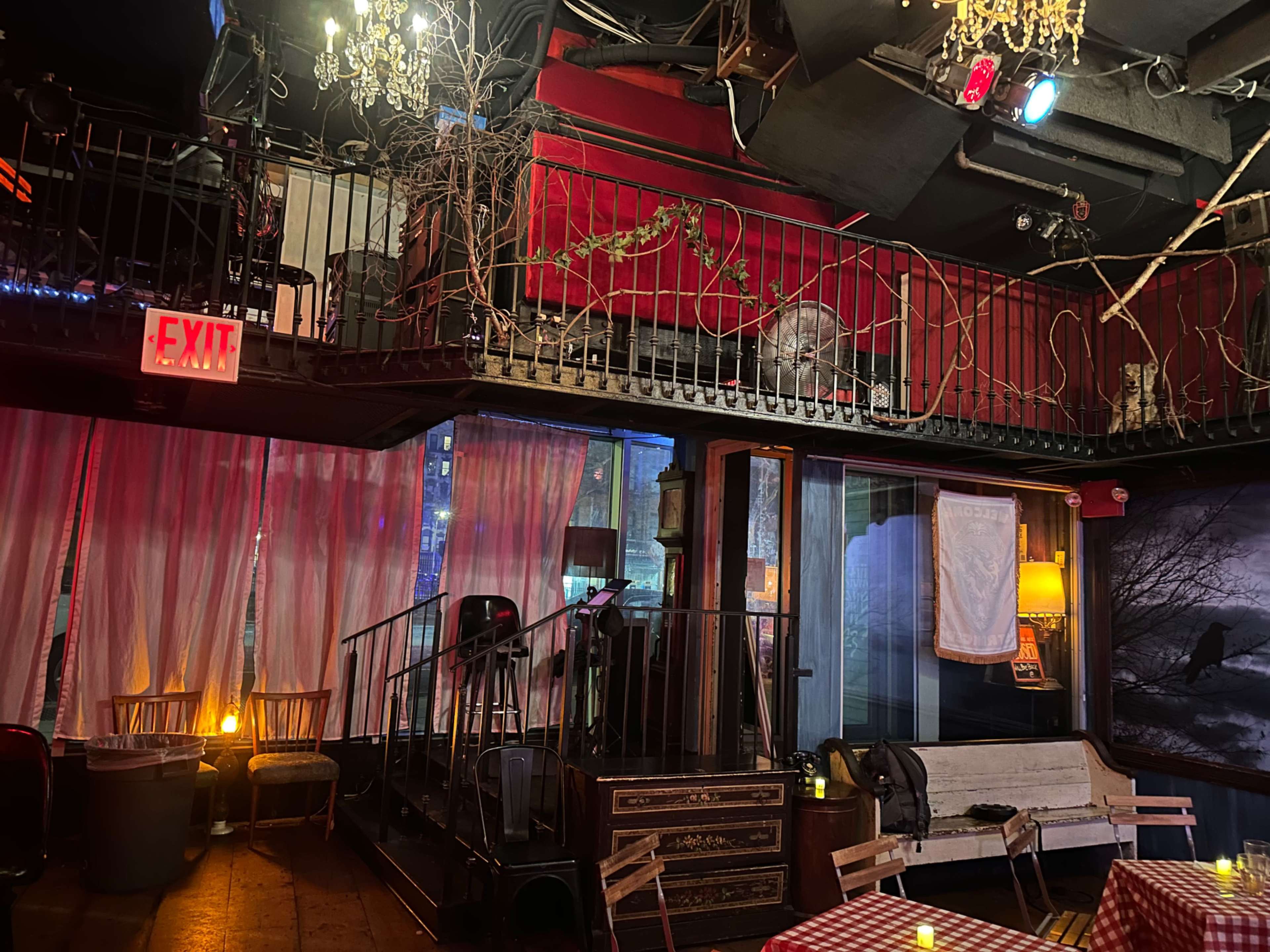 The image shows a dimly lit restaurant interior featuring a balcony area, rustic furniture, and decorative elements like chandeliers and curtains.