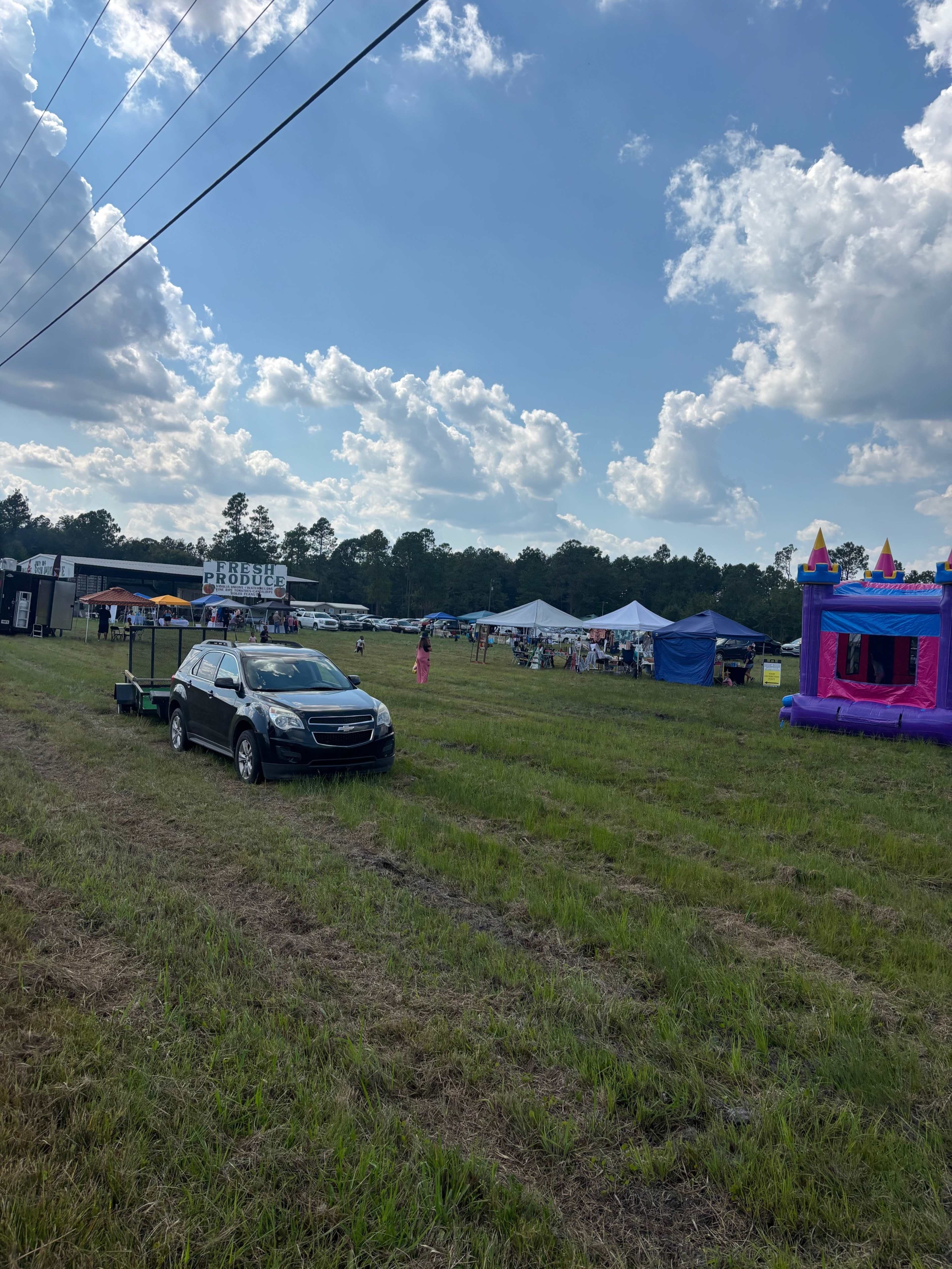 A parked car is visible in a grassy field near several tents and a bounce house, with clouds in the sky above.