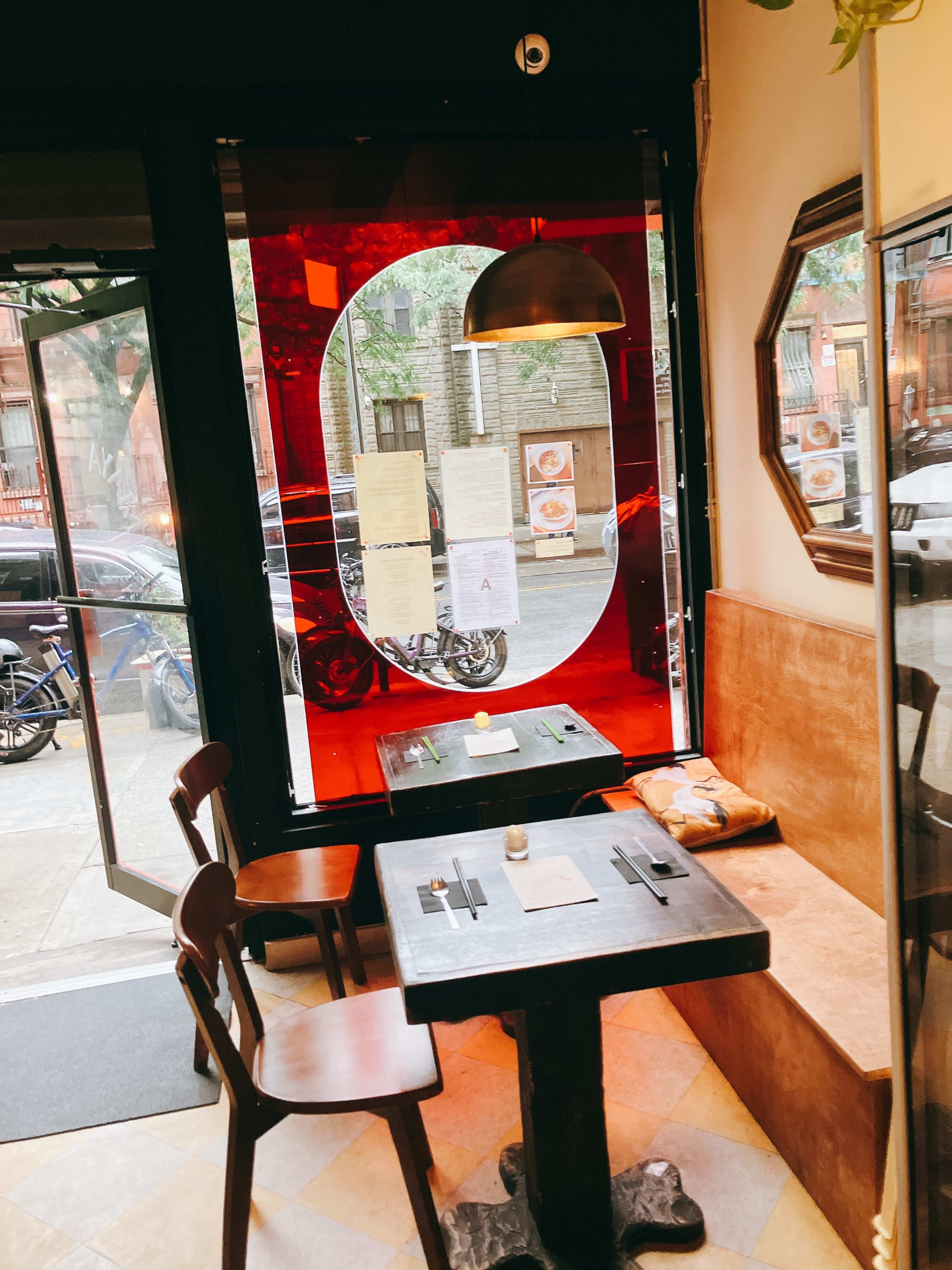 A small restaurant interior features a wooden table and chairs, with a large window that displays a view of the street outside.