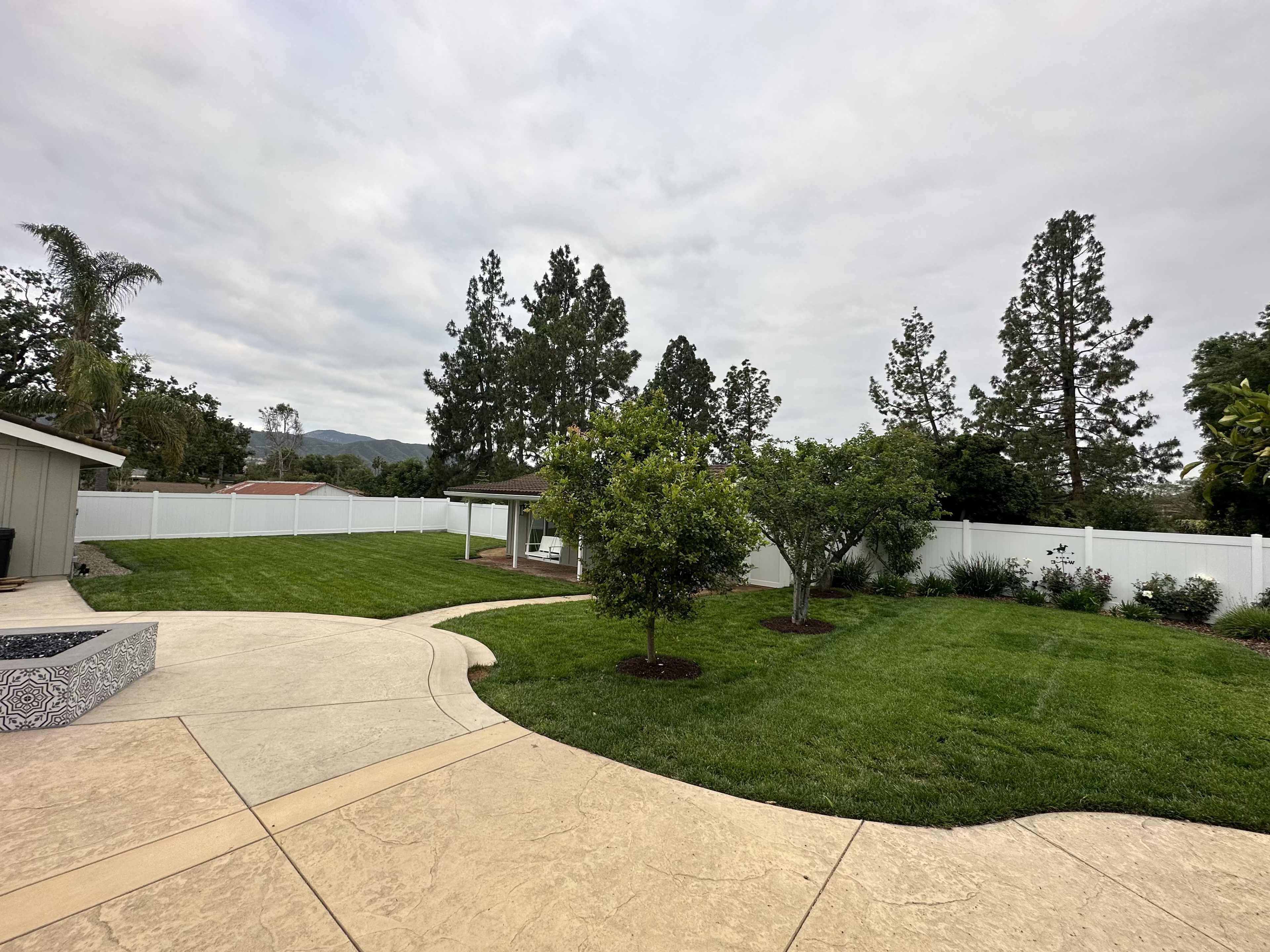 The image shows a well-maintained backyard featuring a neatly mowed lawn, two small trees, a gazebo, and a white fence surrounding the area.