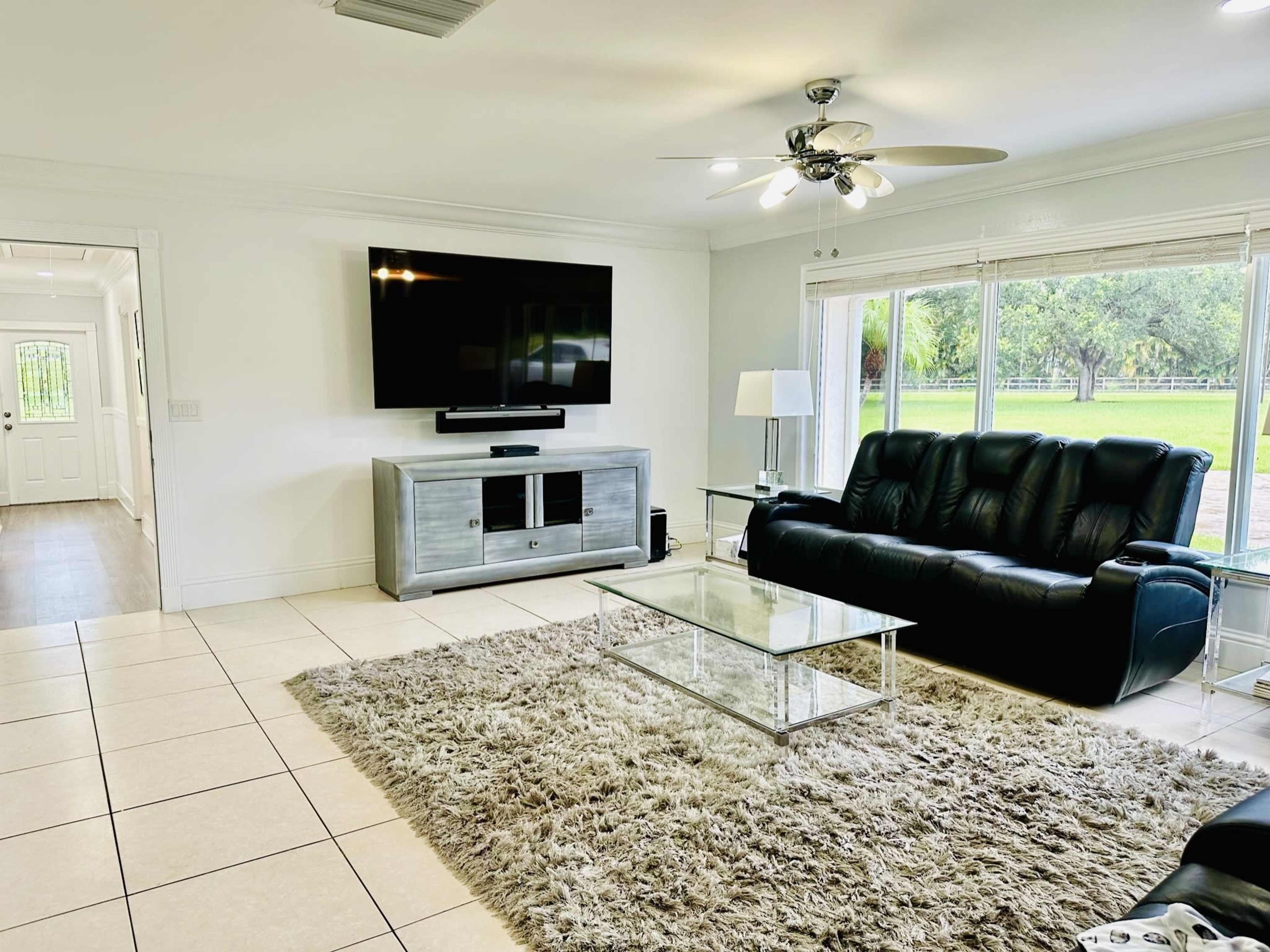 The living room features a black leather sofa, a television mounted on the wall, glass coffee table, and large windows that overlook a grassy area outside.