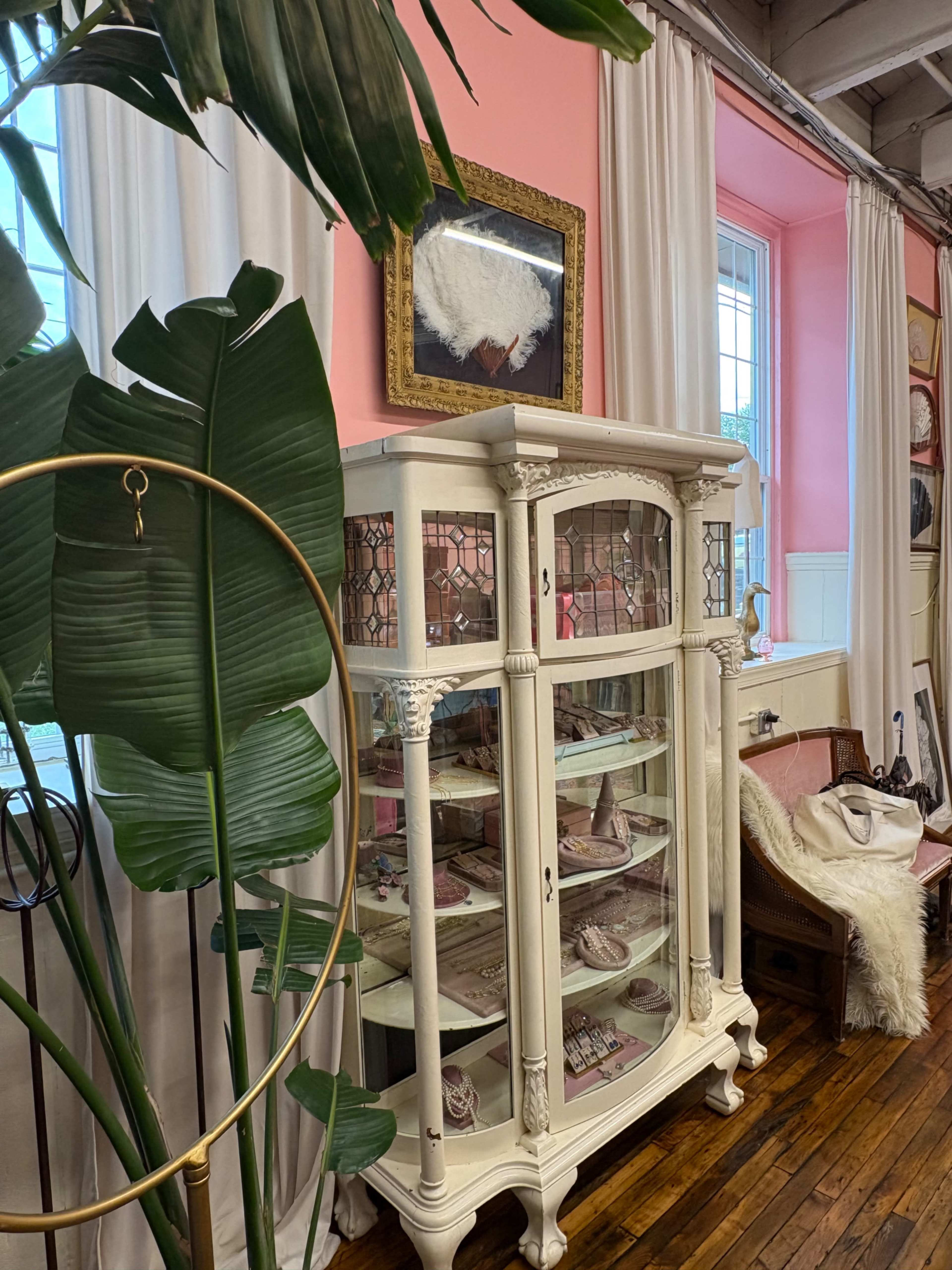 The image shows a vintage white cabinet with glass doors displaying various items, positioned against a pink wall with curtains and a decorative painting above it.