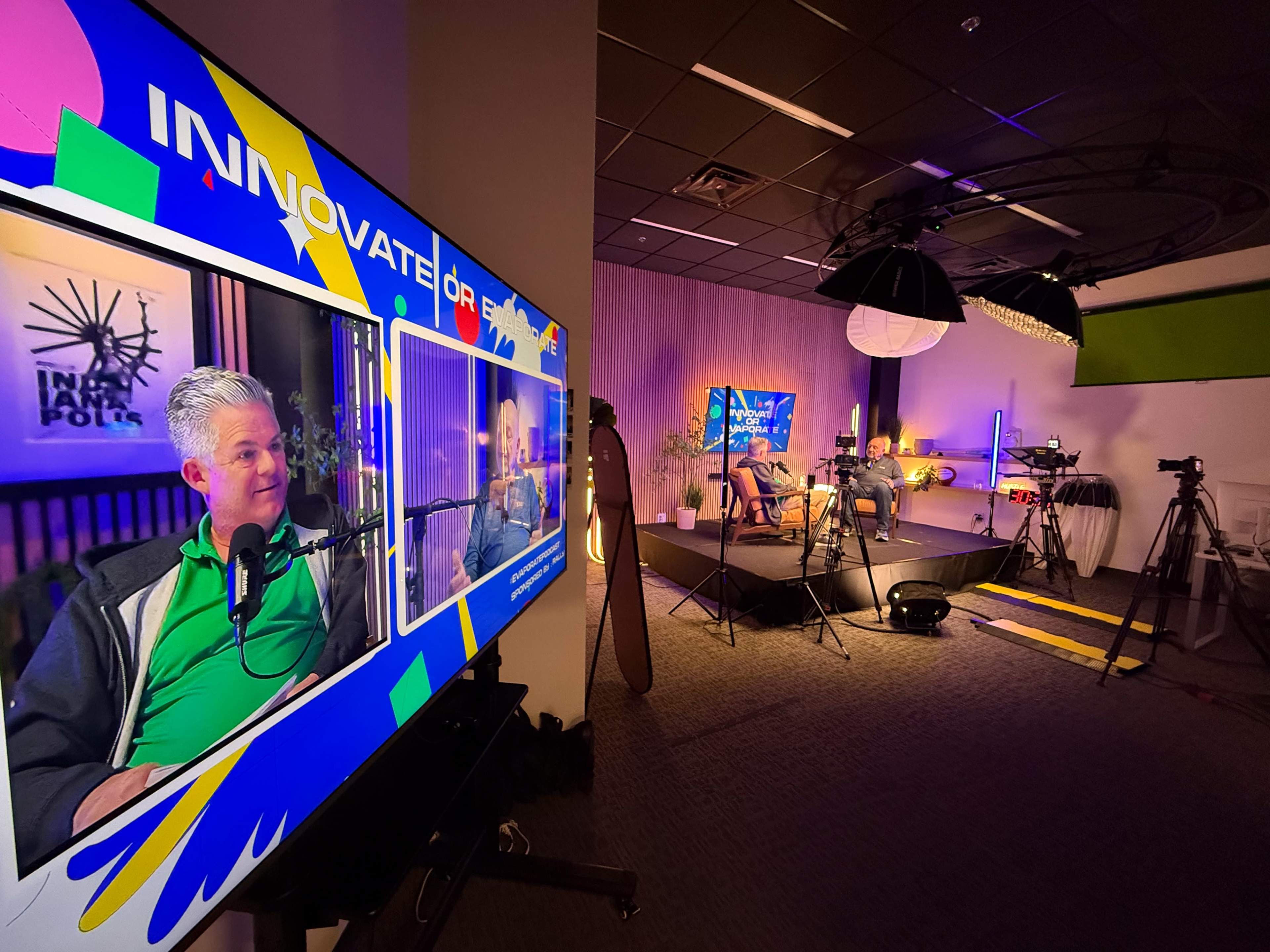 The image shows a recording studio setup with multiple cameras, a large screen displaying a podcast host, and a brightly lit background featuring colorful decor.