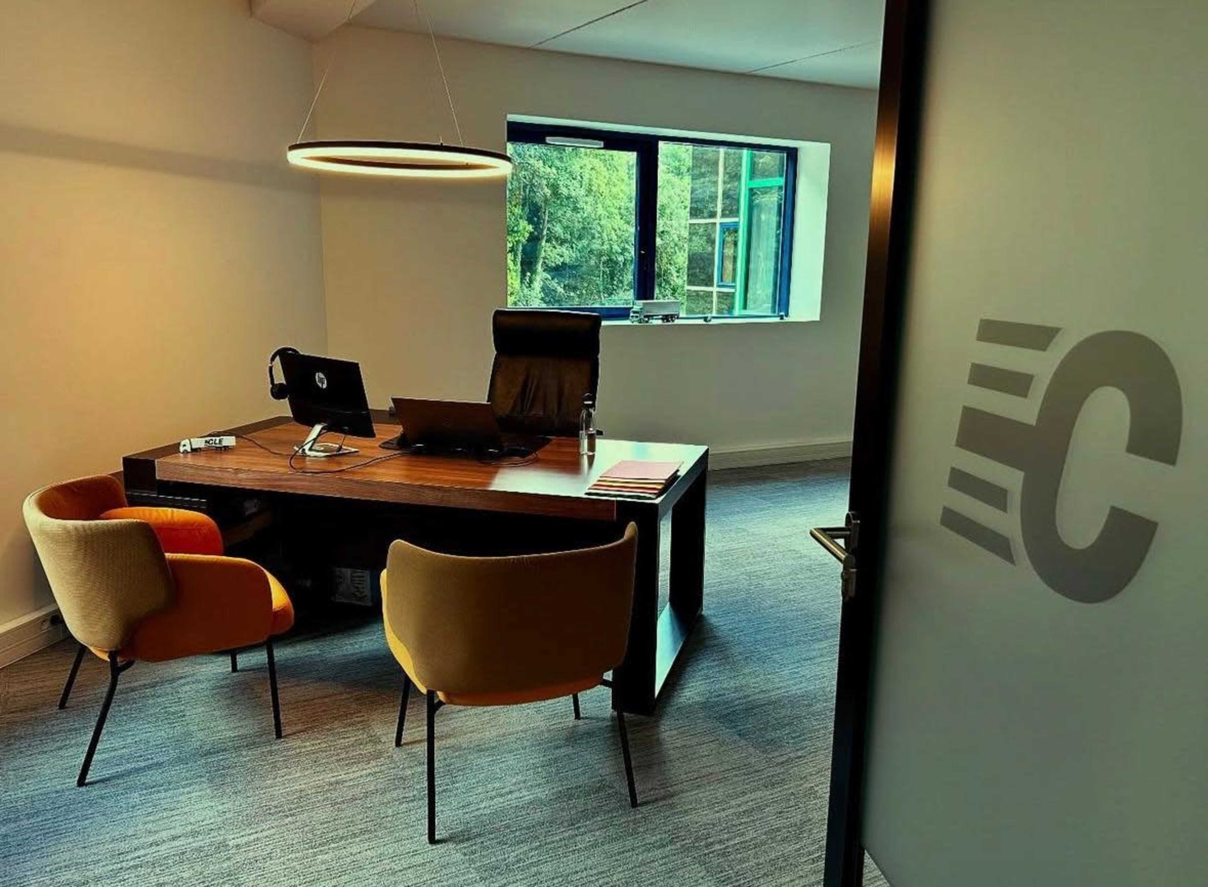 The image shows a modern office interior featuring a wooden desk with a computer, two chairs, and large windows allowing natural light.