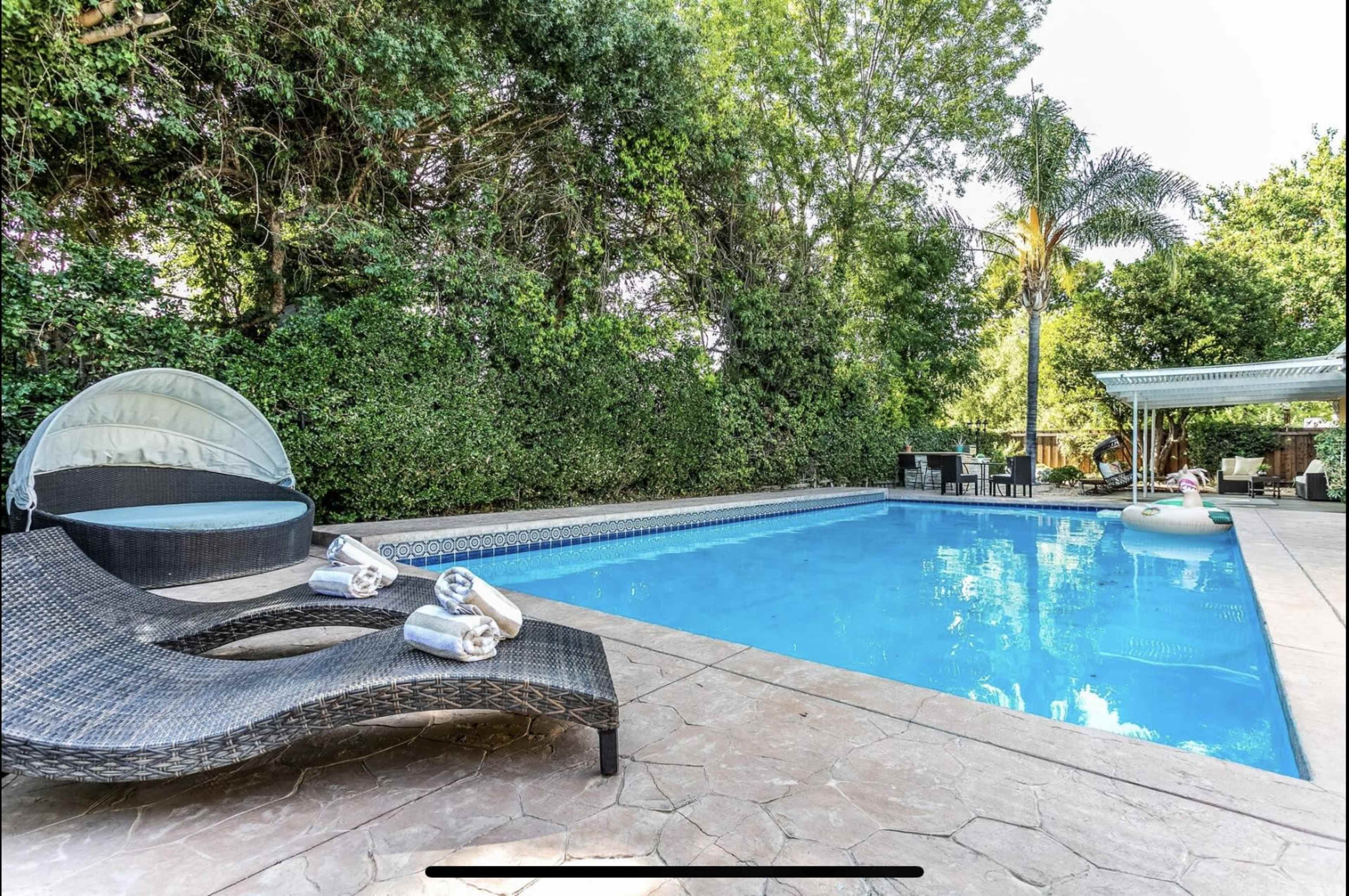 A swimming pool with a lounge area, surrounded by greenery and patio furniture, is partially shaded by trees.