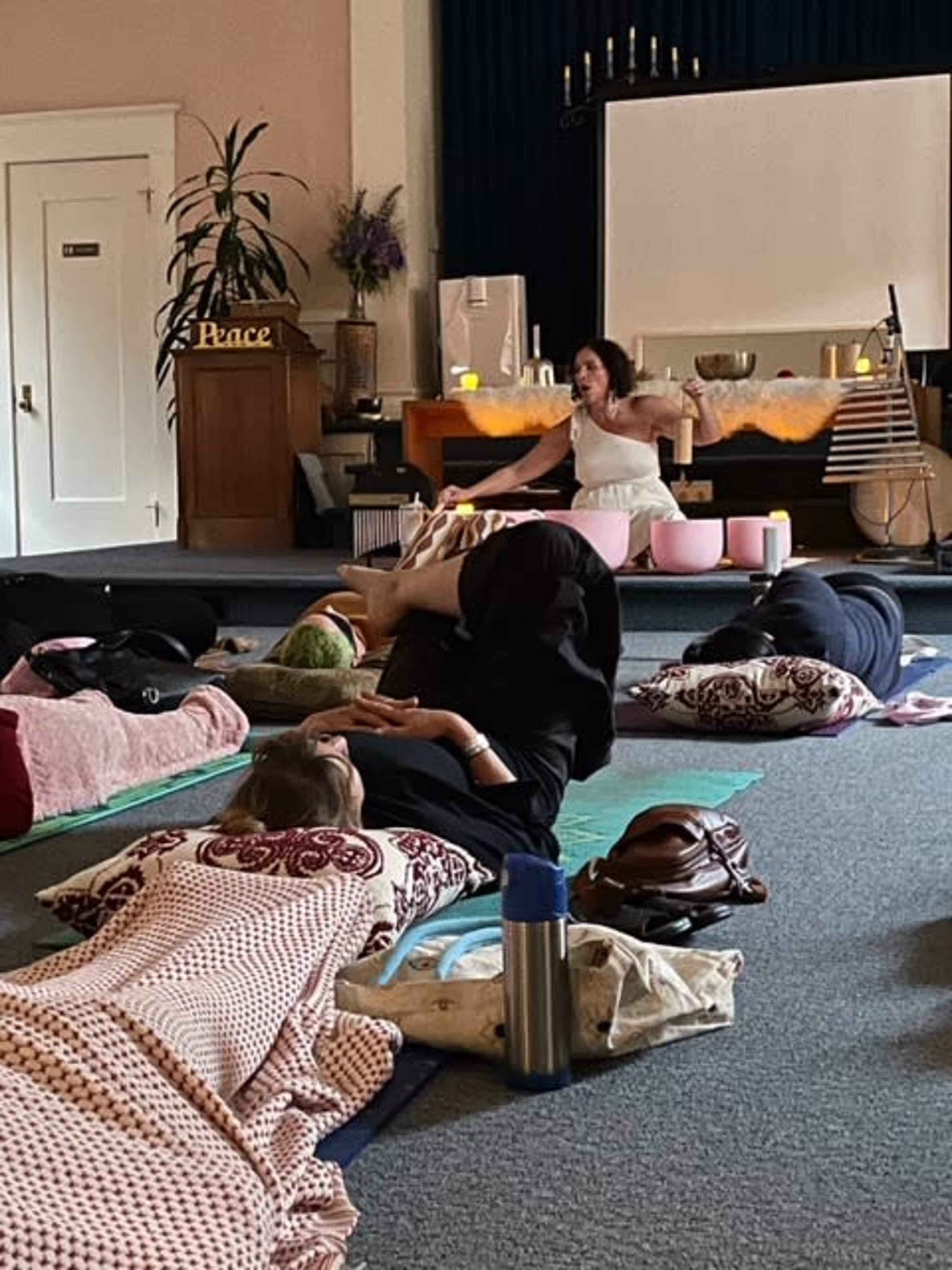 A group of people is lying on mats in a room while a woman addresses them from the front, surrounded by pillows and soft lighting.