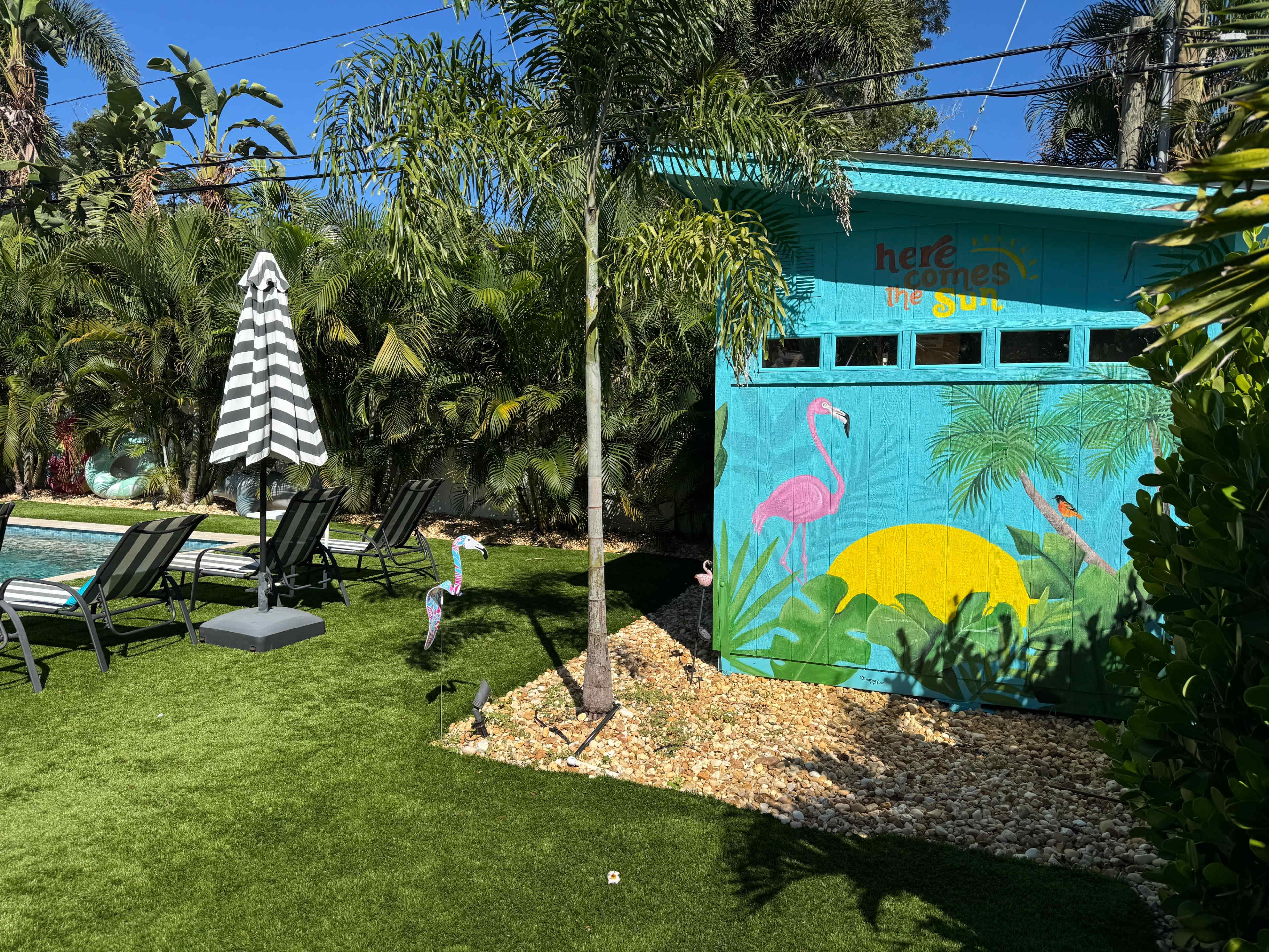 The image shows a vibrant outdoor area featuring a swimming pool, lounge chairs, a striped umbrella, and a mural depicting flamingos and tropical scenery on a shed.