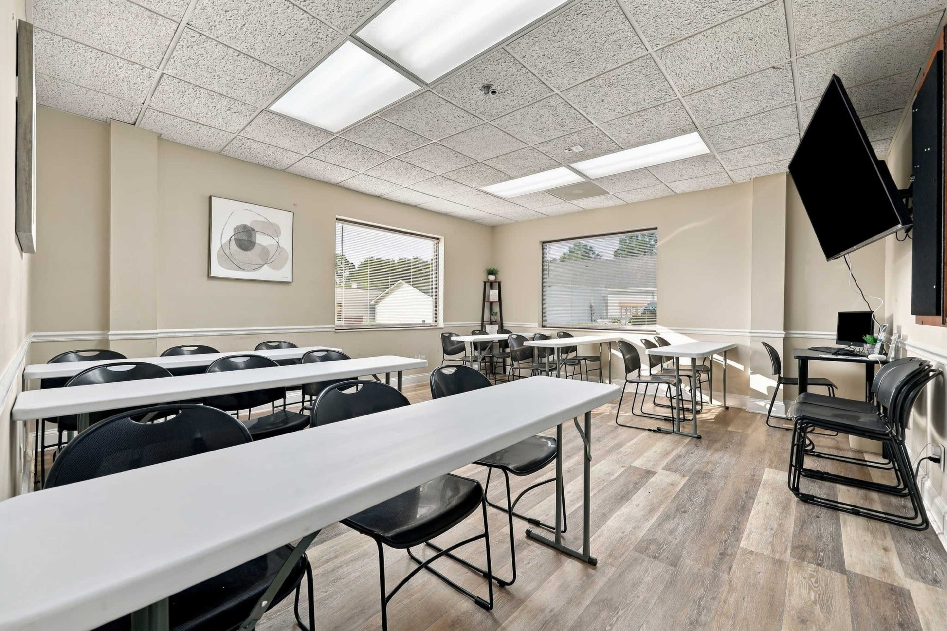 The image shows a bright, empty classroom with rows of tables and chairs arranged for a meeting or class, along with a television mounted on the wall.