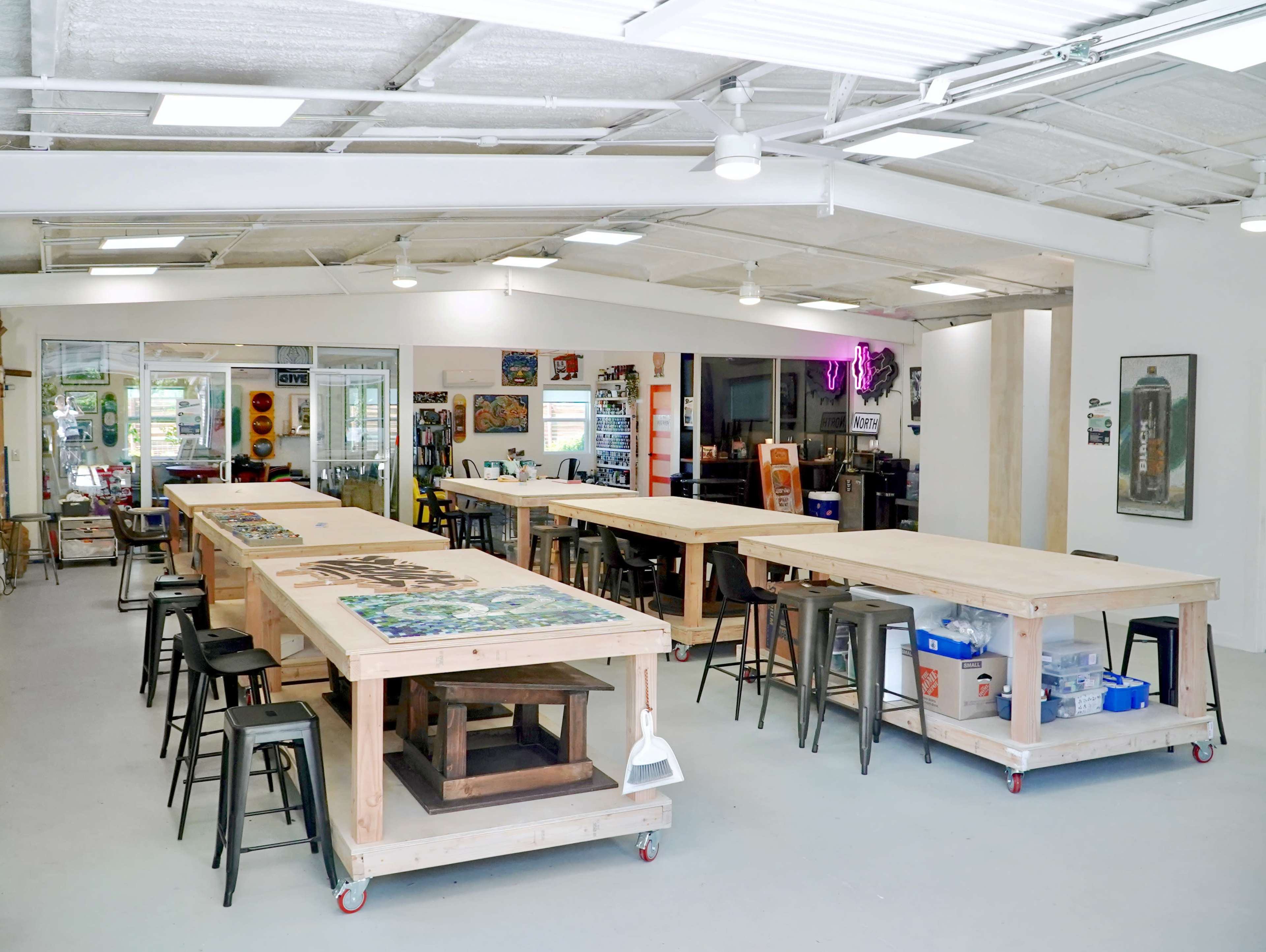 The image shows a spacious workshop with multiple large, wooden tables and metal stools arranged in a bright, industrial-style room.