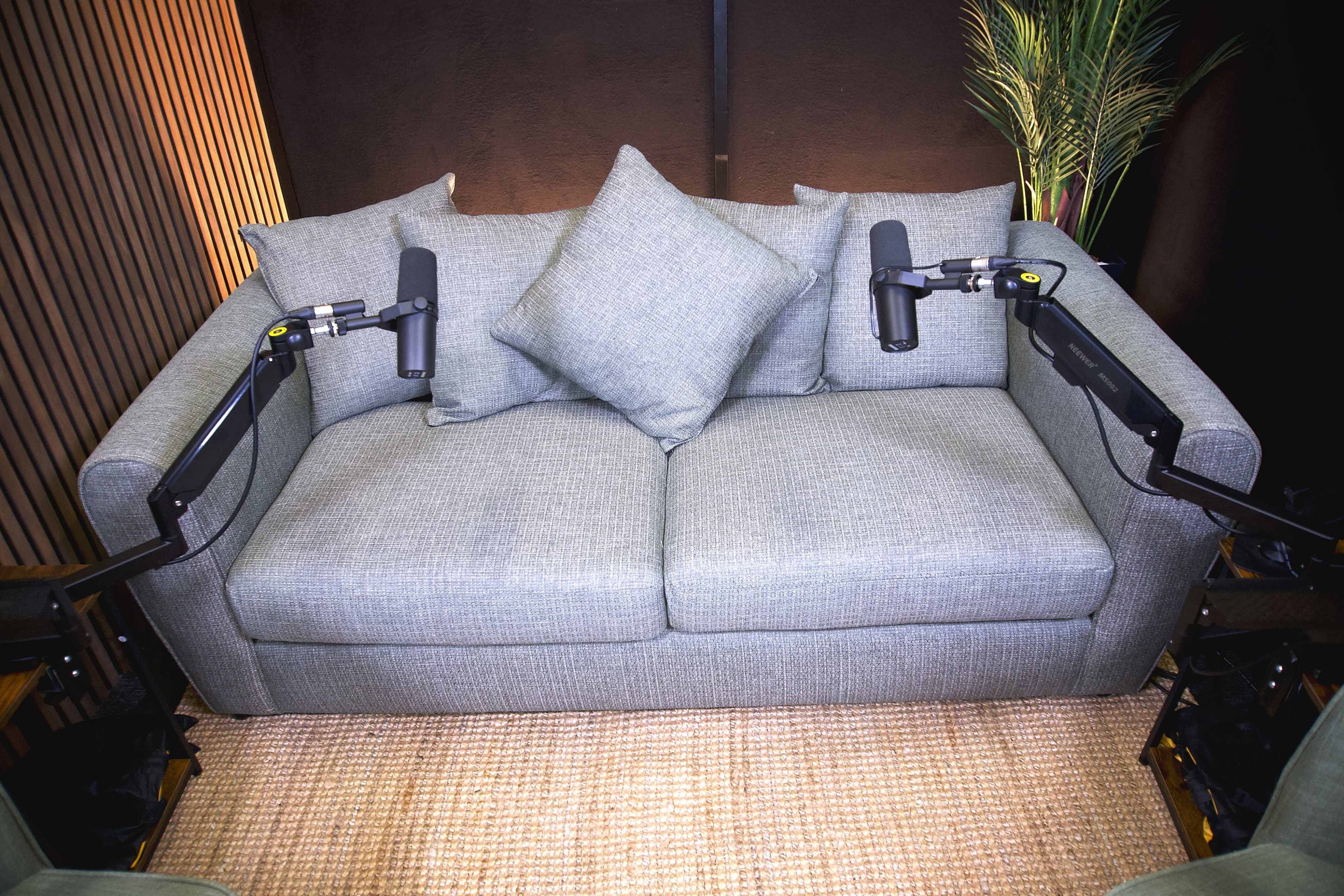 A gray upholstered couch with two cushions is positioned in a room, flanked by two microphones on adjustable arms.