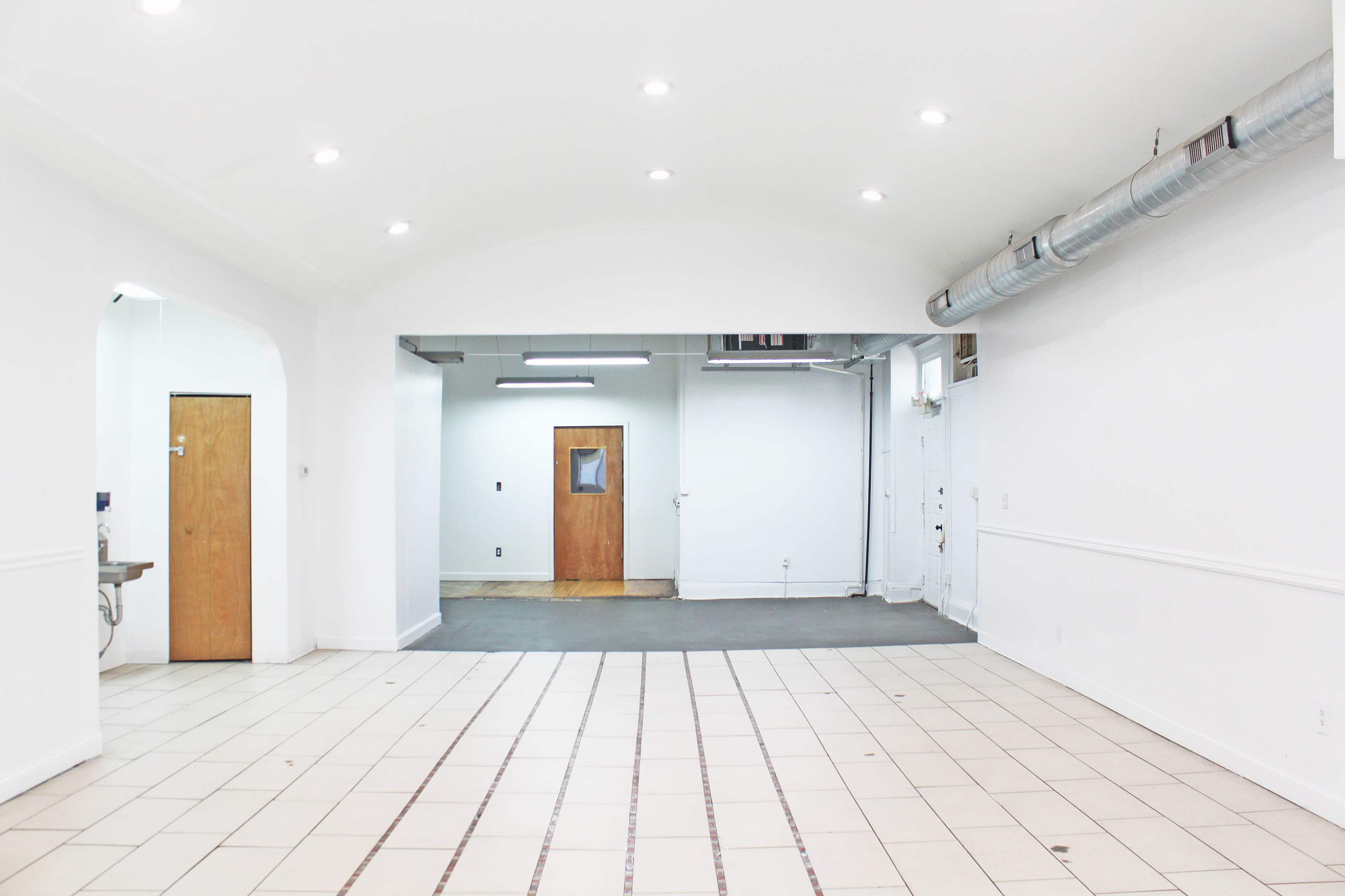 The image shows a spacious, empty room with white walls and tiled flooring, featuring two doorways and exposed ductwork on the ceiling.