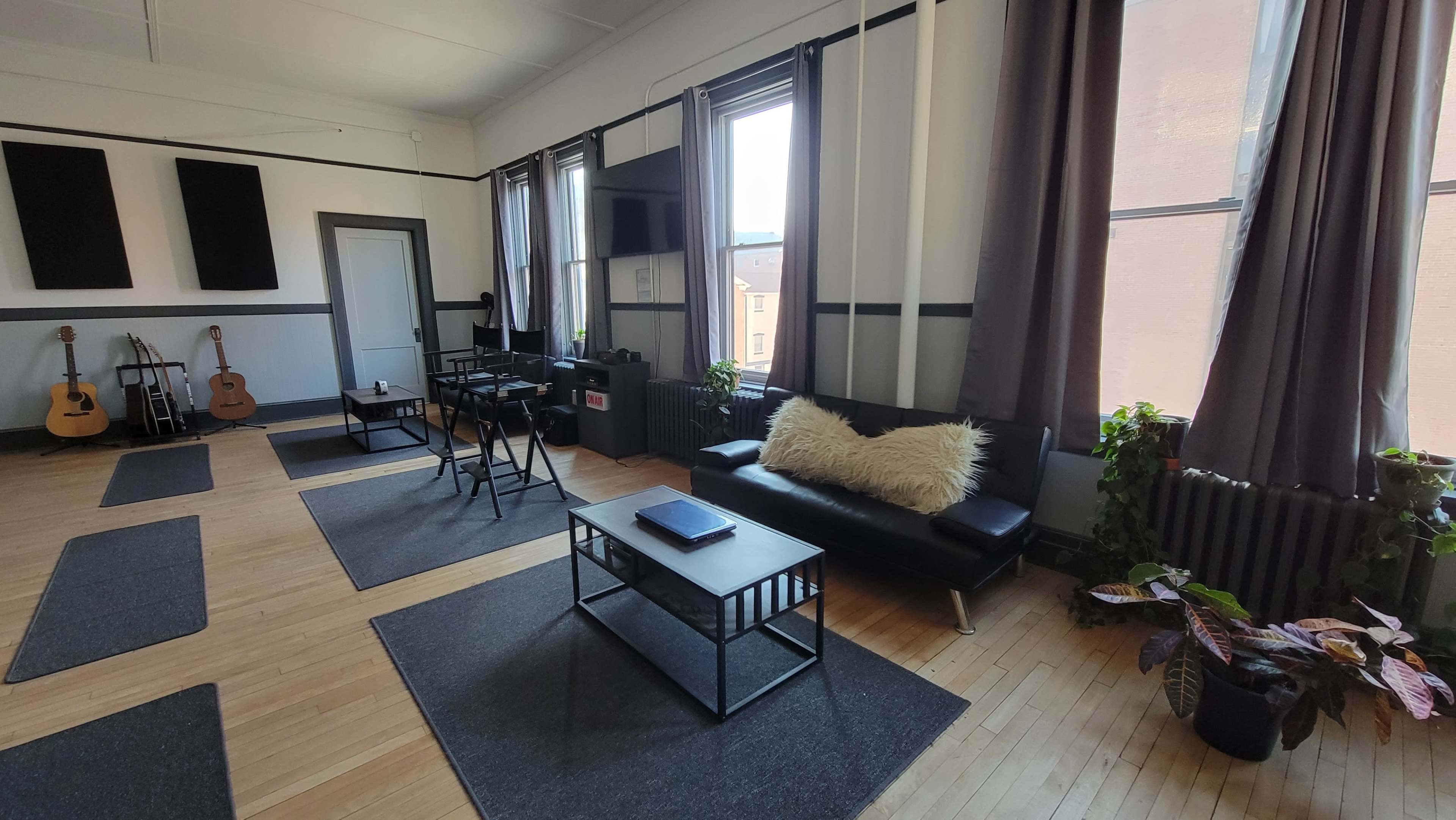 A bright, open room with wooden floors, large windows, and minimal furniture including a black sofa, two tables, and potted plants.