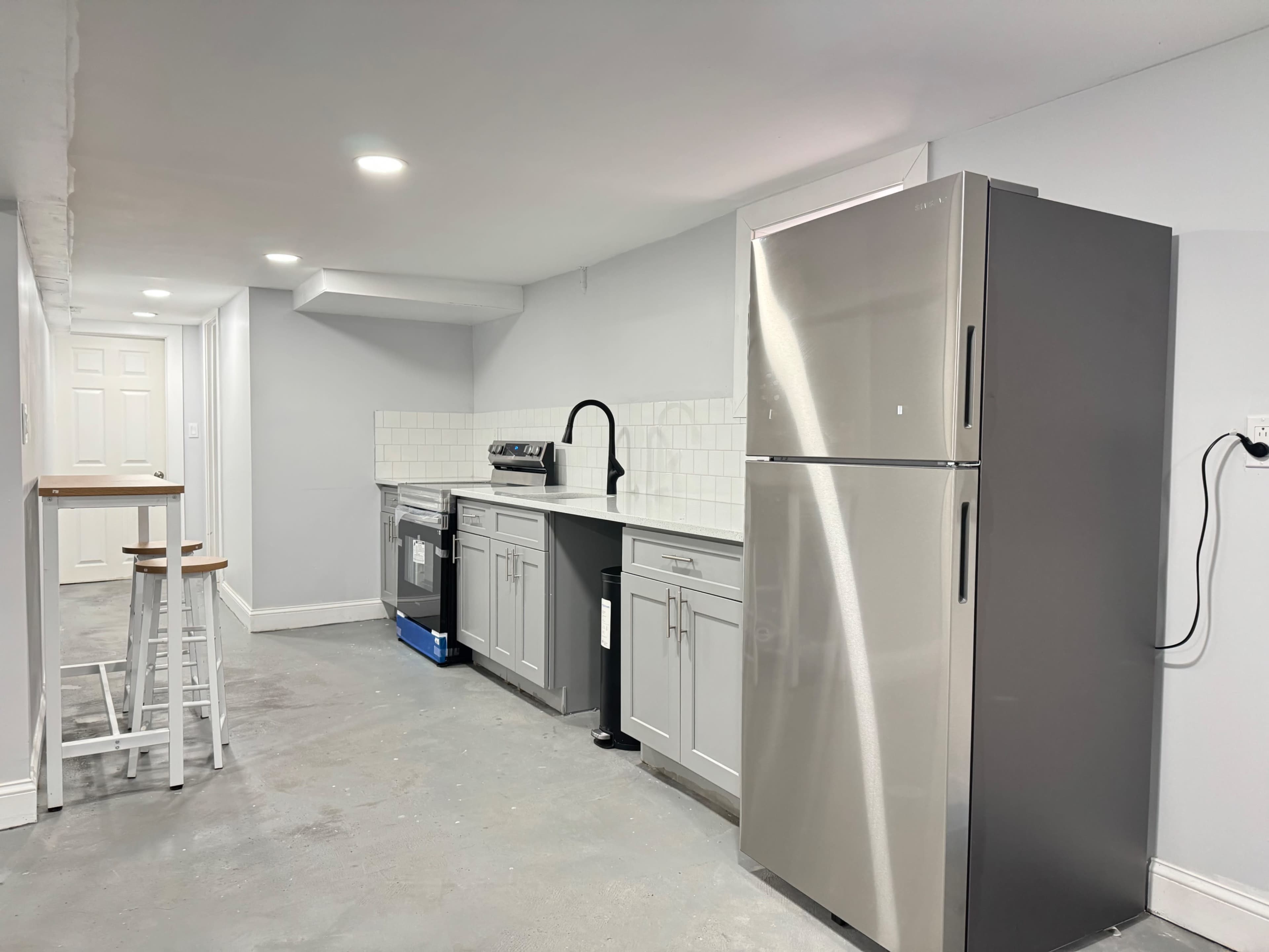 A modern kitchen area with gray cabinets, a stainless steel refrigerator, and a small table with stools against the wall.