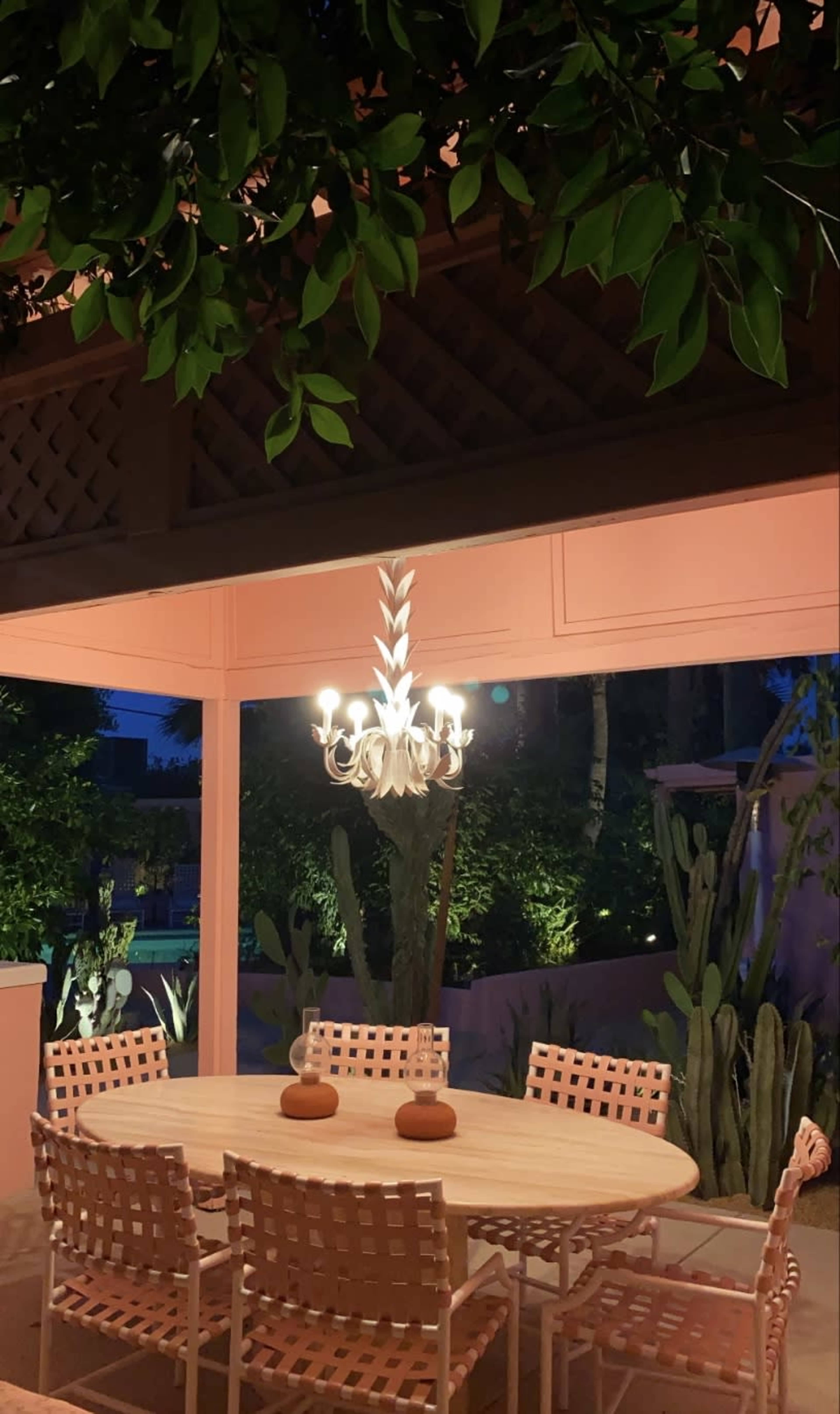 A round dining table with six chairs is set under a chandelier in a shaded outdoor space surrounded by greenery.