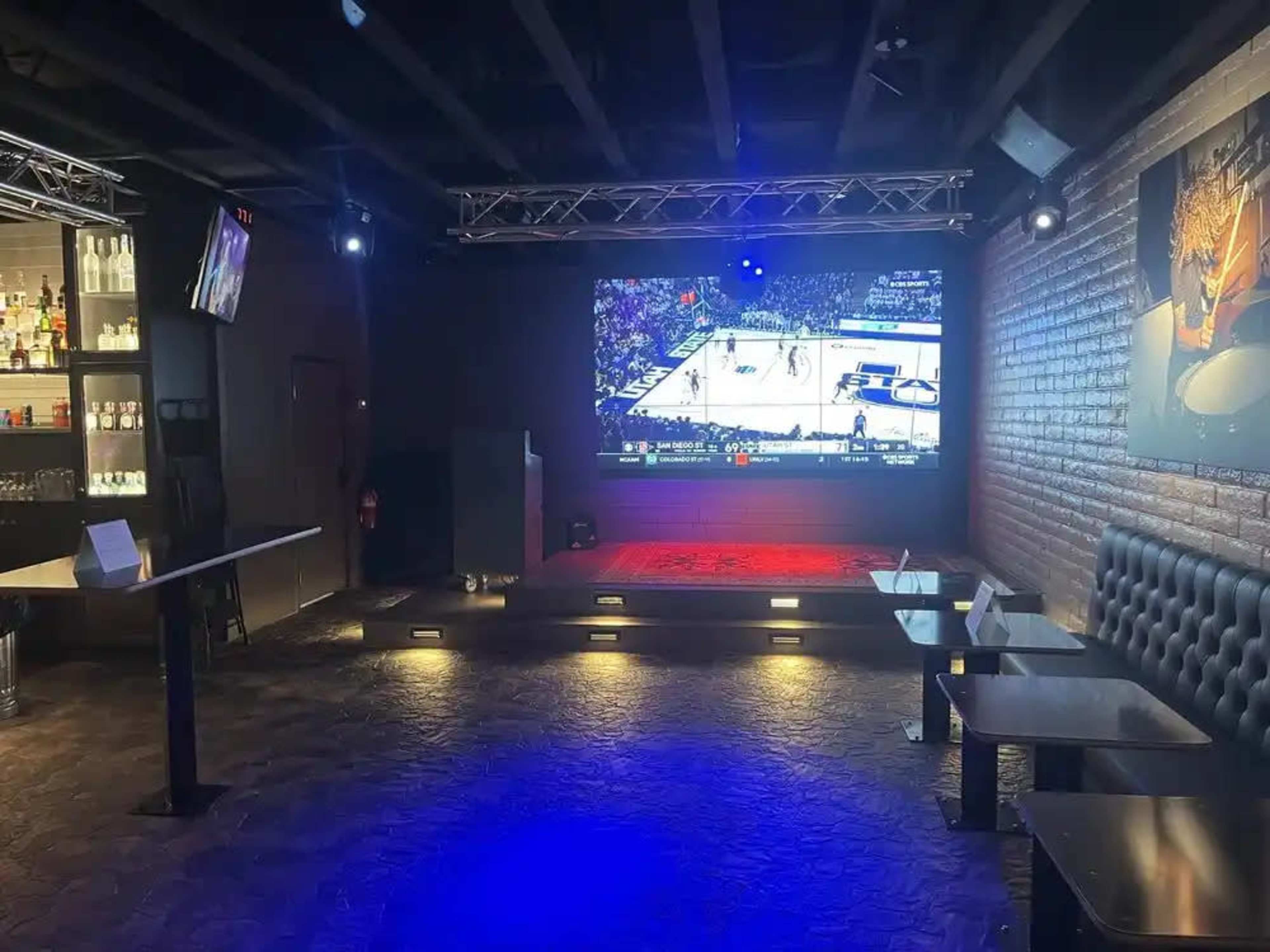The image shows a bar interior featuring a large screen displaying a basketball game, with seating areas and a well-stocked back bar illuminated under dim lighting.