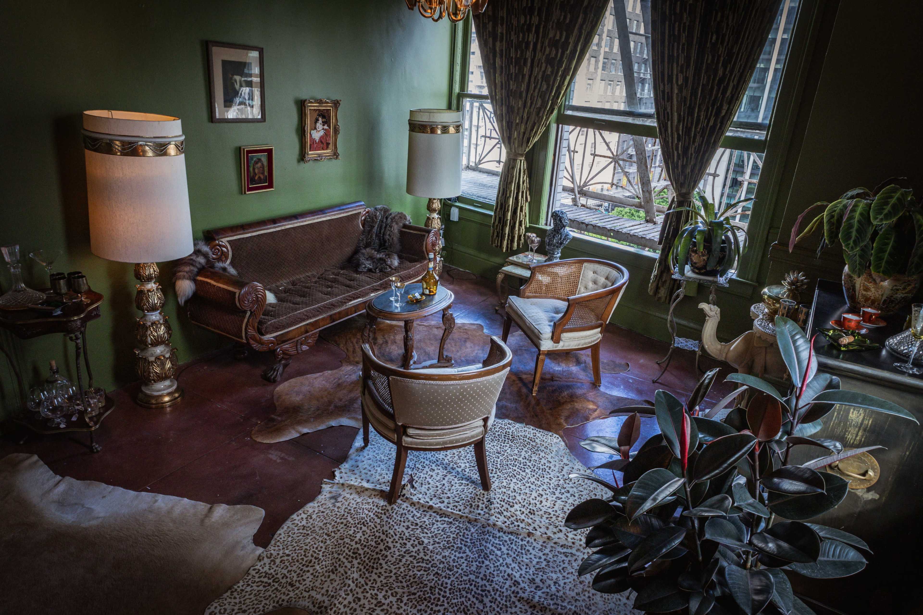 The image shows a vintage-styled living room with a green wall, a brown sofa, two upholstered chairs, and large windows allowing natural light to enter.