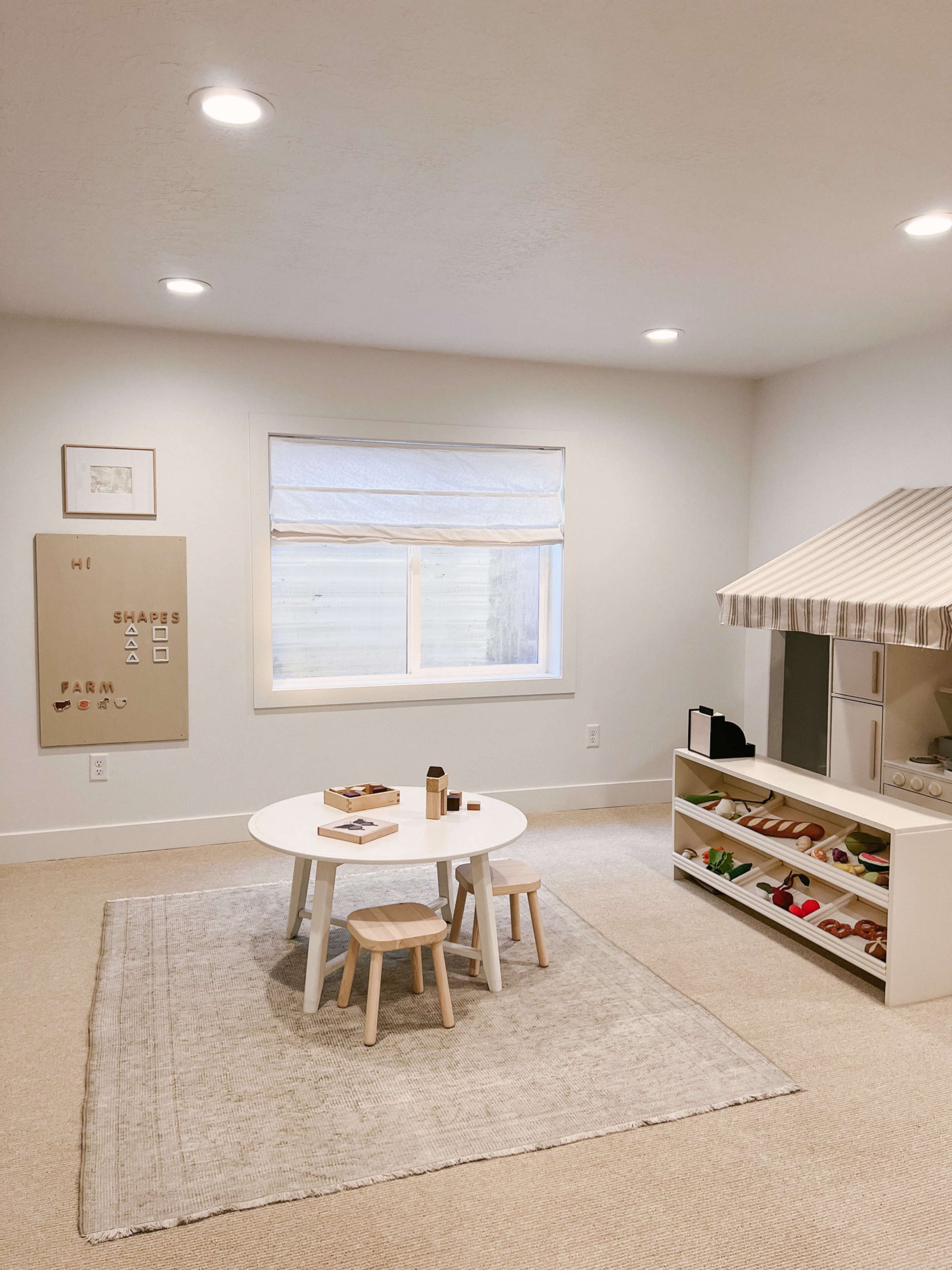 A minimalist playroom with a circular table, two small stools, and a shelf filled with toys, featuring a plush rug and a window with a light-filtering treatment.