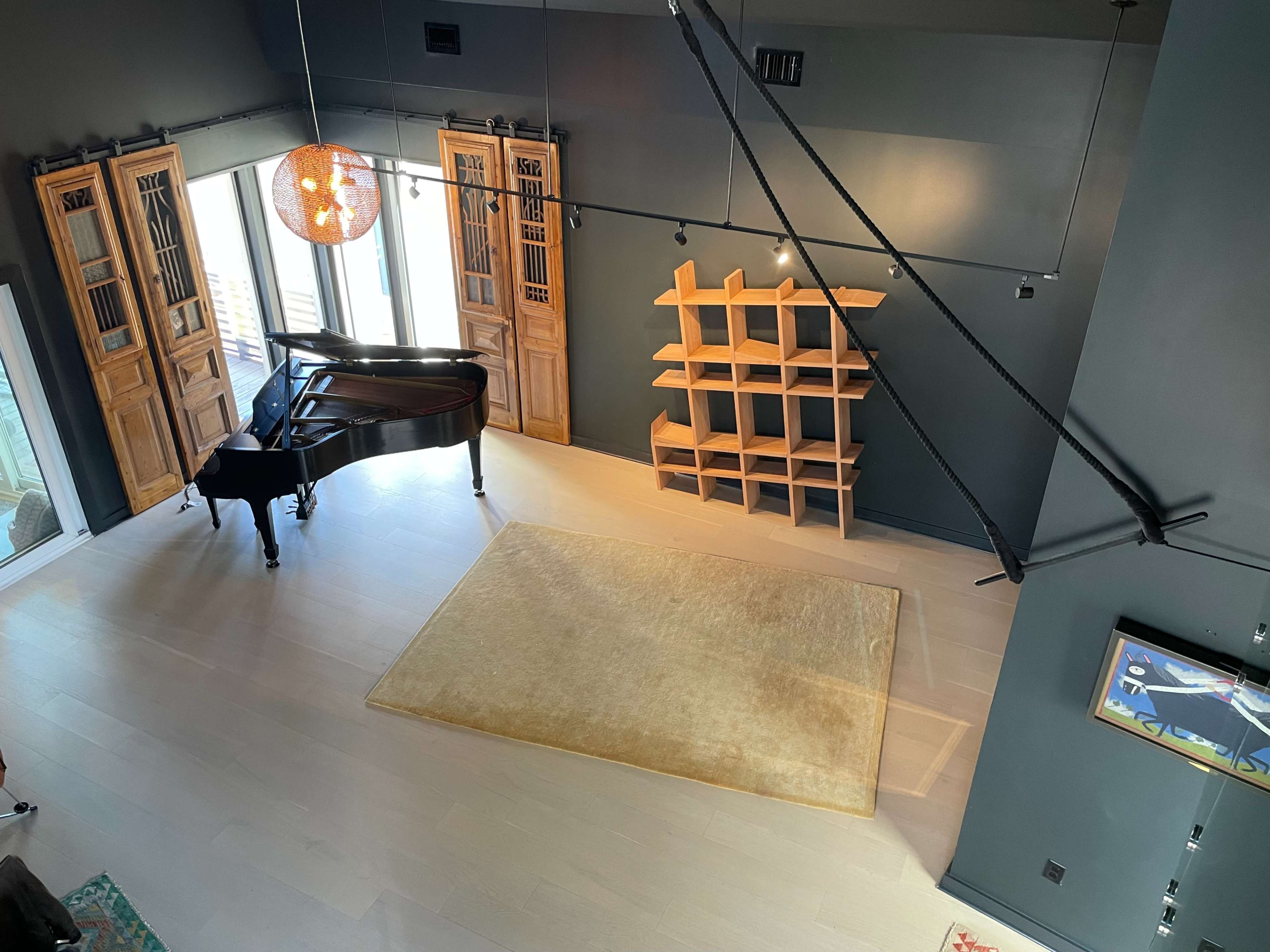The image shows a spacious room with a grand piano, wooden storage units, and a large area rug on a light wooden floor, illuminated by a pendant light hanging from the ceiling.