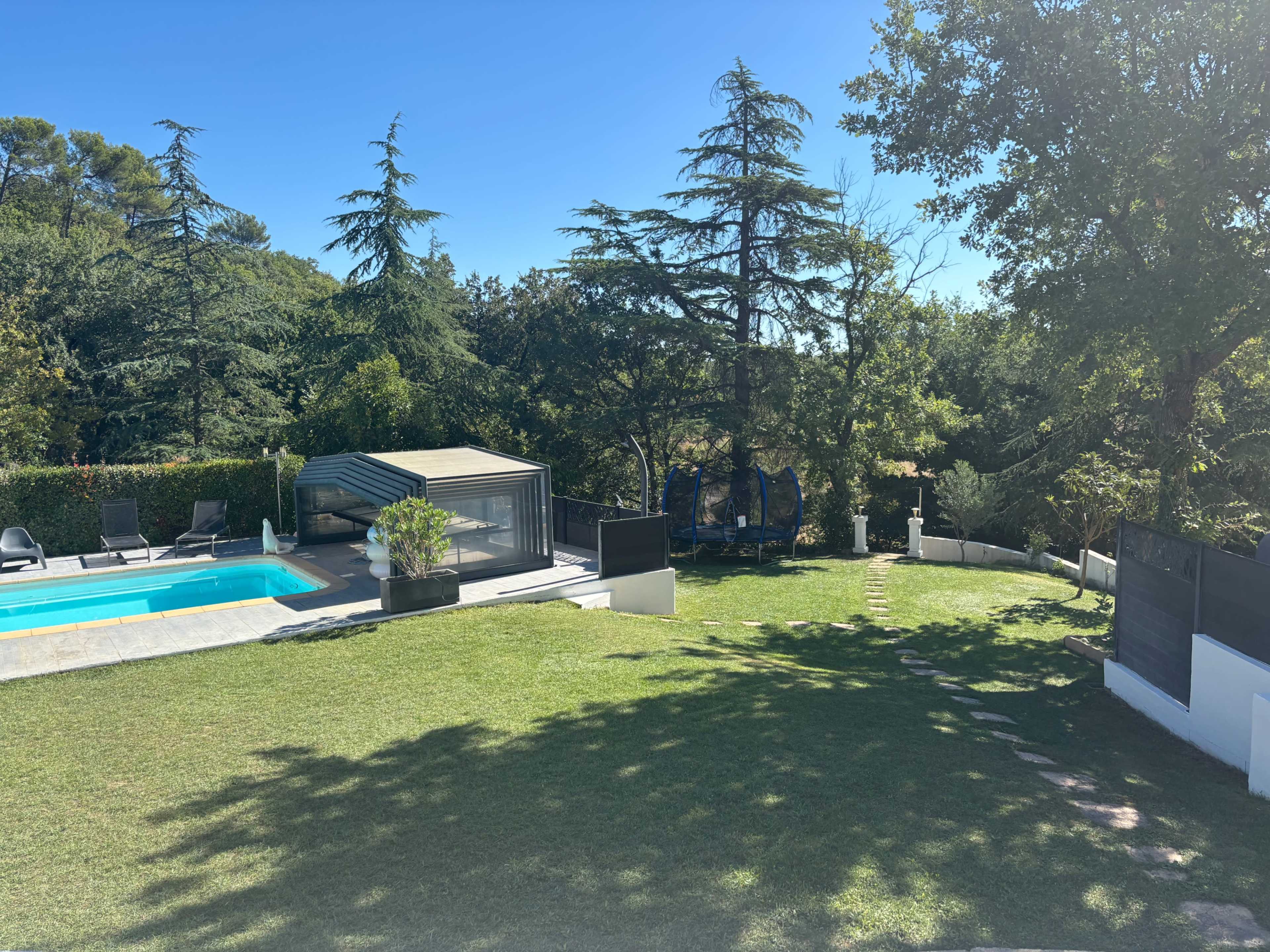 The image shows a backyard featuring a swimming pool, a modern glass structure, and a lawn with trees and lounge chairs.