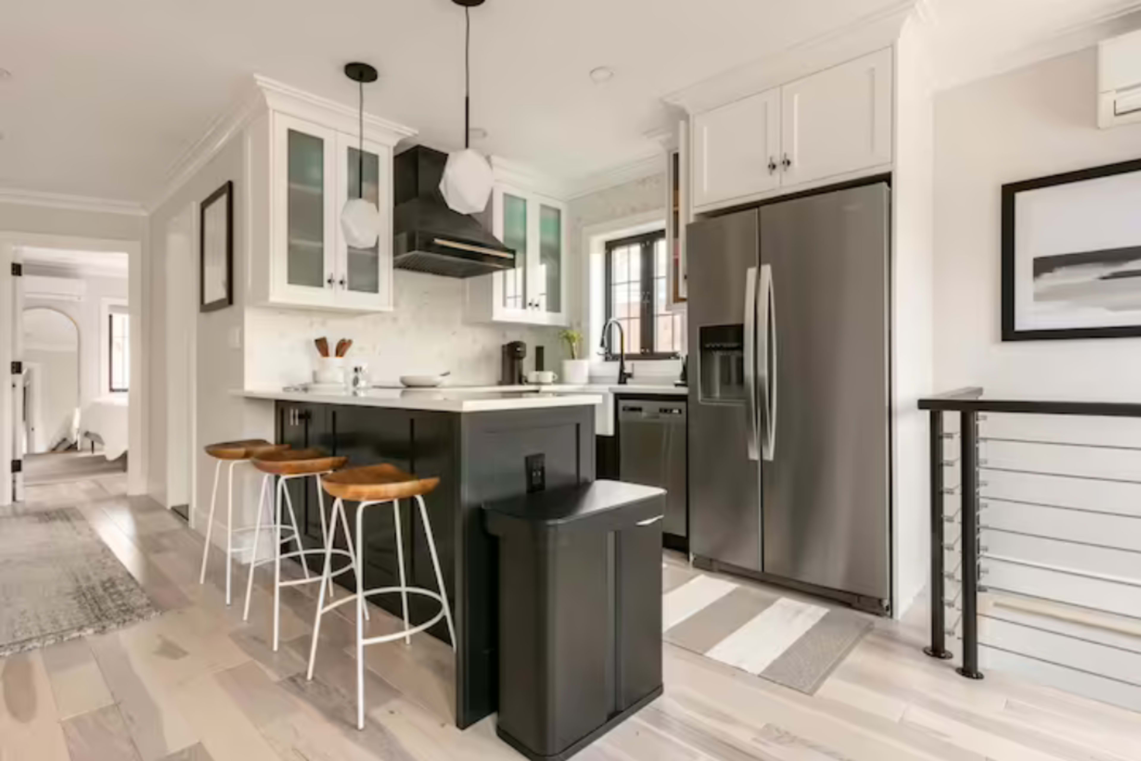 A modern kitchen features a central island with three stools, stainless steel appliances, and cabinetry with glass accents.