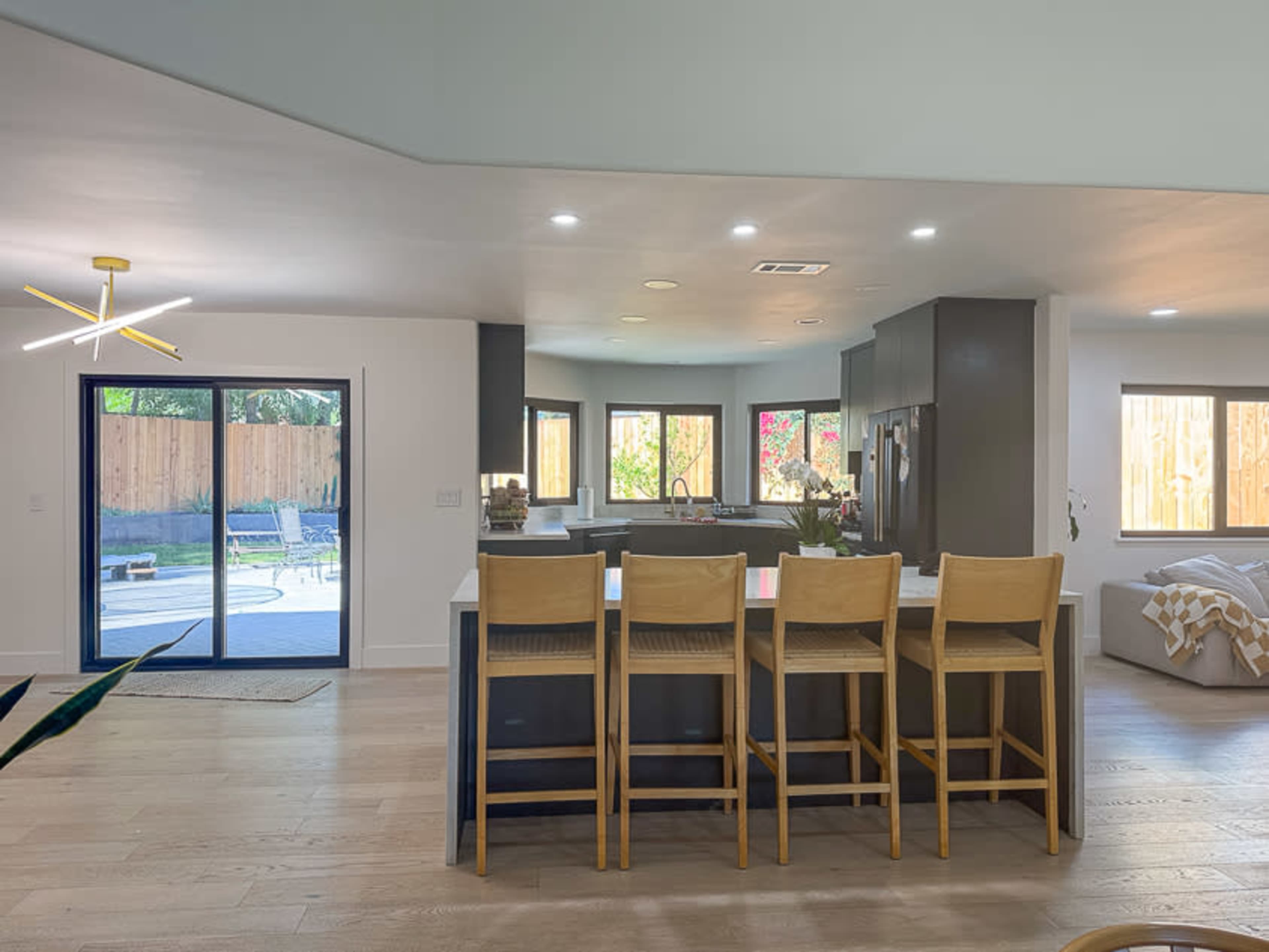 The image shows a modern kitchen with four wooden bar stools facing a kitchen island, large windows allowing natural light, and a view of an outdoor patio.