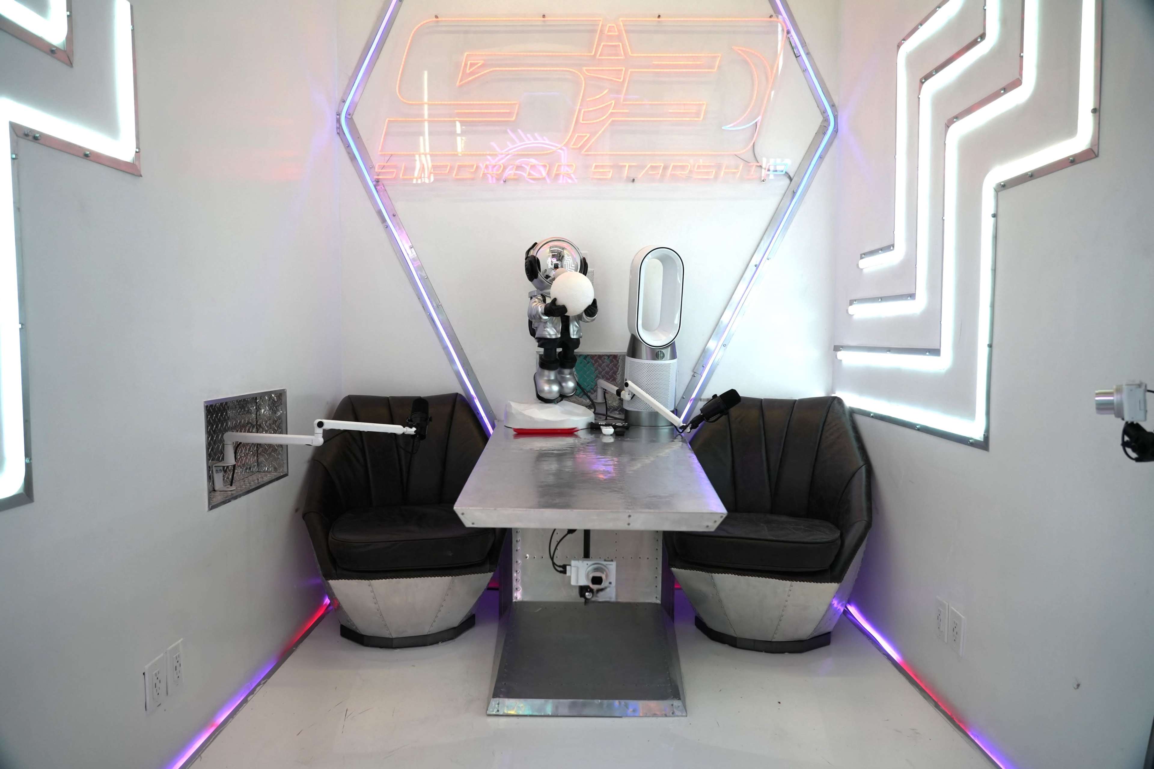 A futuristic booth with two cushioned chairs, a metallic table, and a robotic figure, surrounded by glowing geometric light fixtures on the walls.