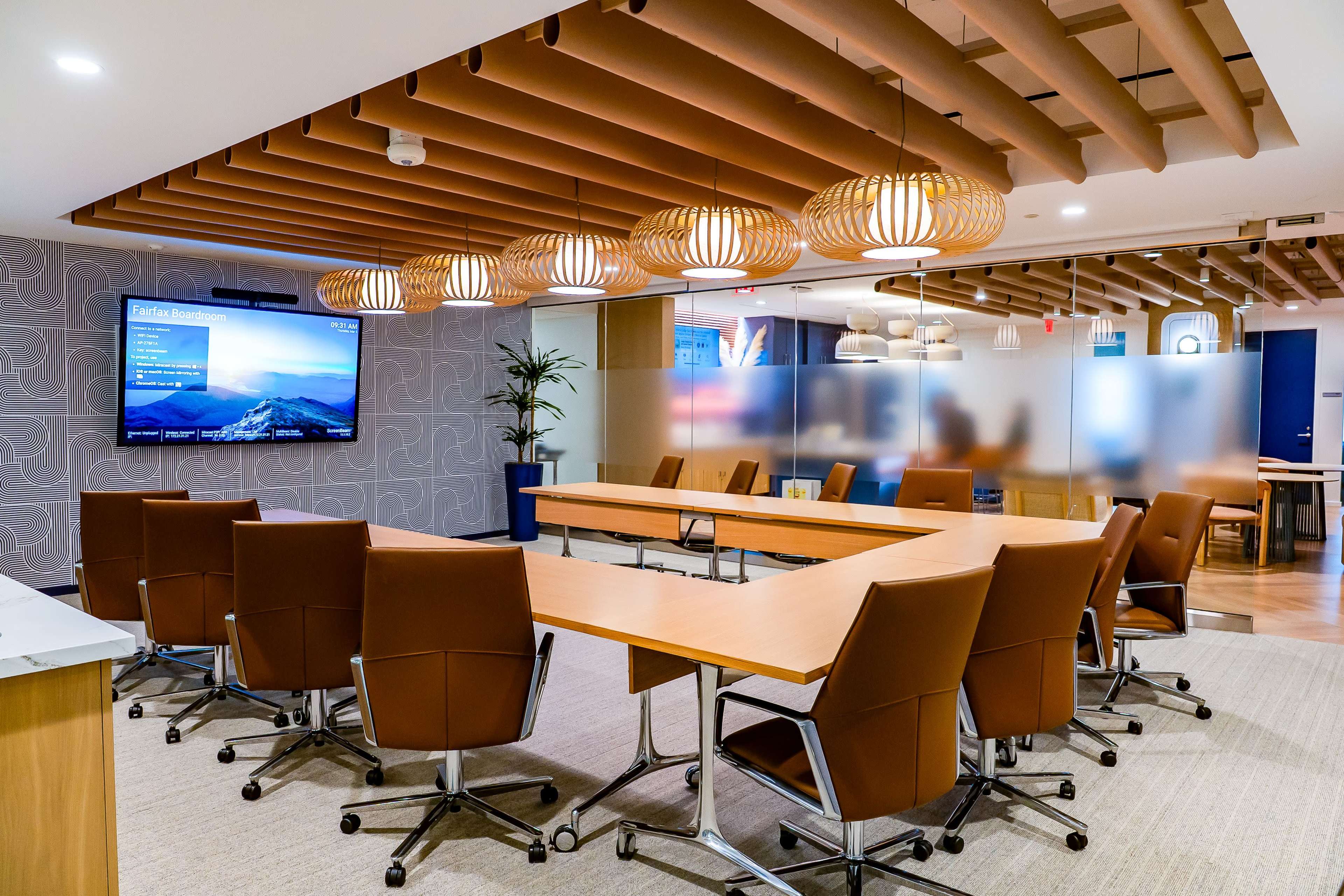 The image shows a modern conference room featuring a large table surrounded by ergonomic chairs, a wall-mounted screen displaying a presentation, and decorative overhead lighting.
