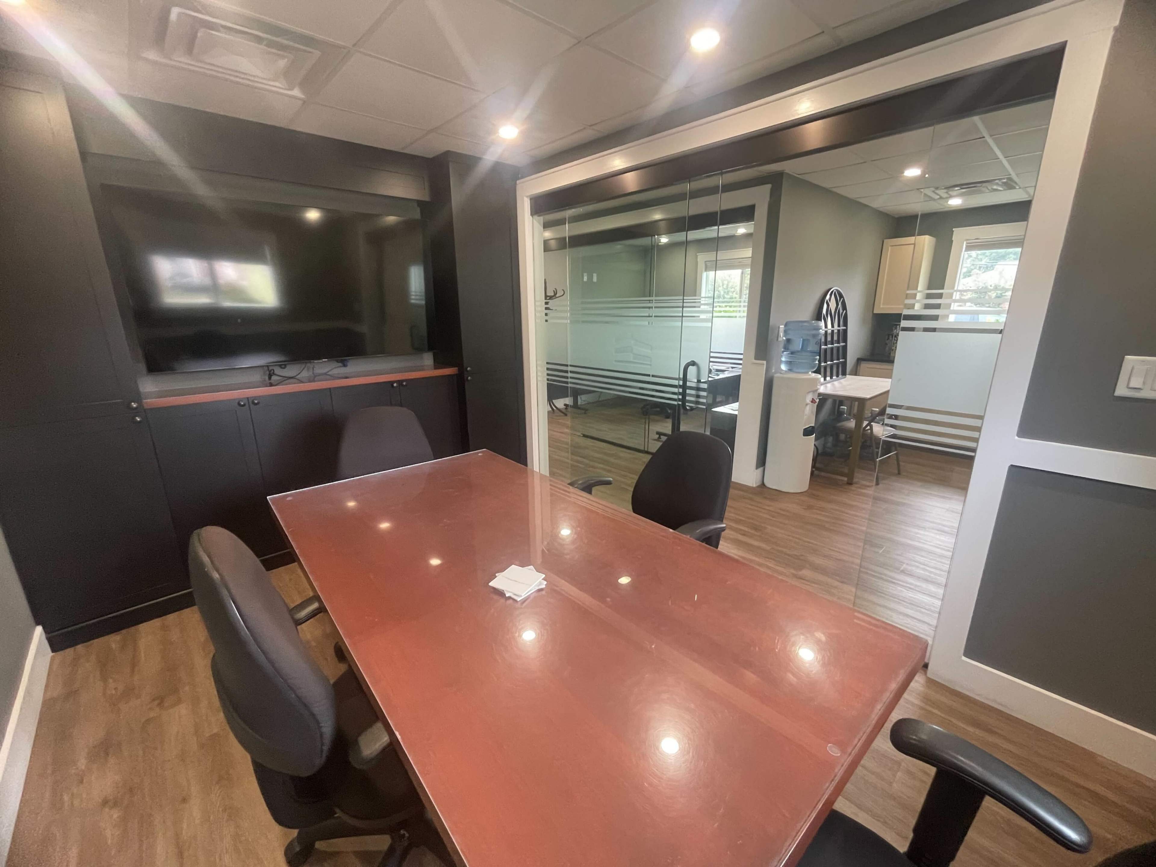 The image shows a modern conference room with a wooden table and black chairs, featuring a glass wall and a large screen on one side.