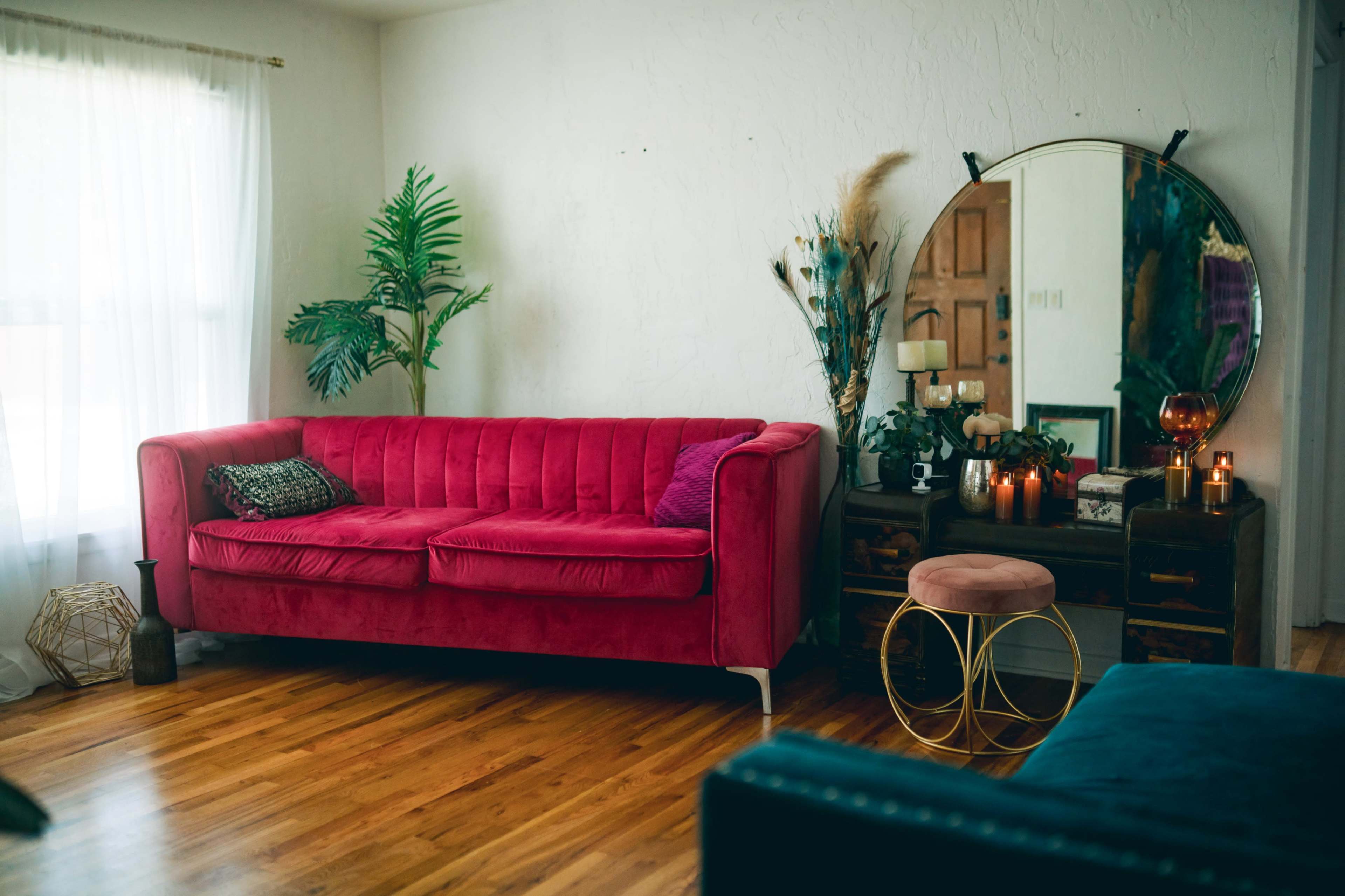 The image shows a stylish living room featuring a pink velvet sofa, a round mirror, a small round stool, and decorative plants.