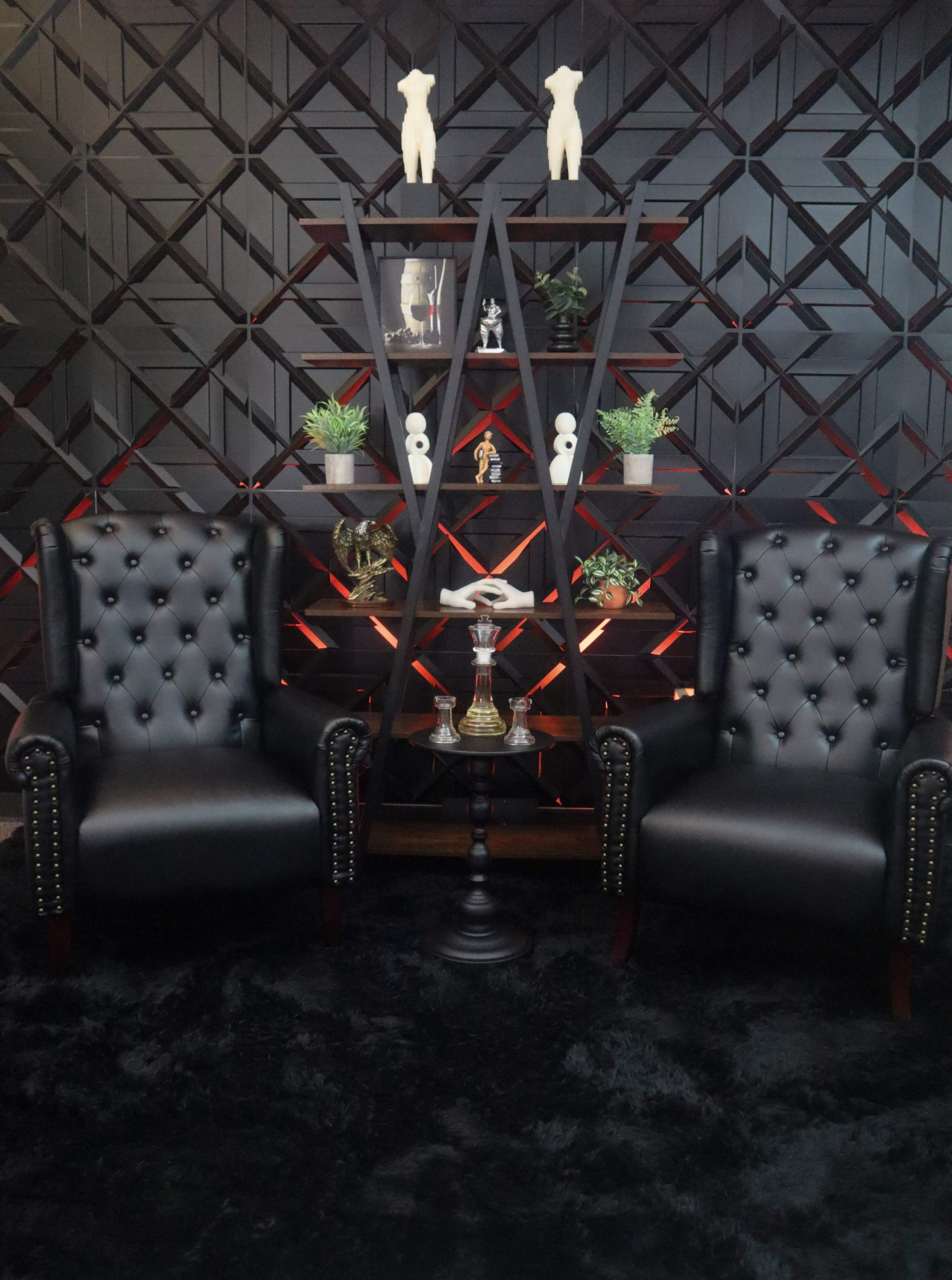 The image features two black tufted chairs positioned on either side of a small table, with a backdrop of a dark geometric patterned wall and a decorative shelf containing various items and plants.