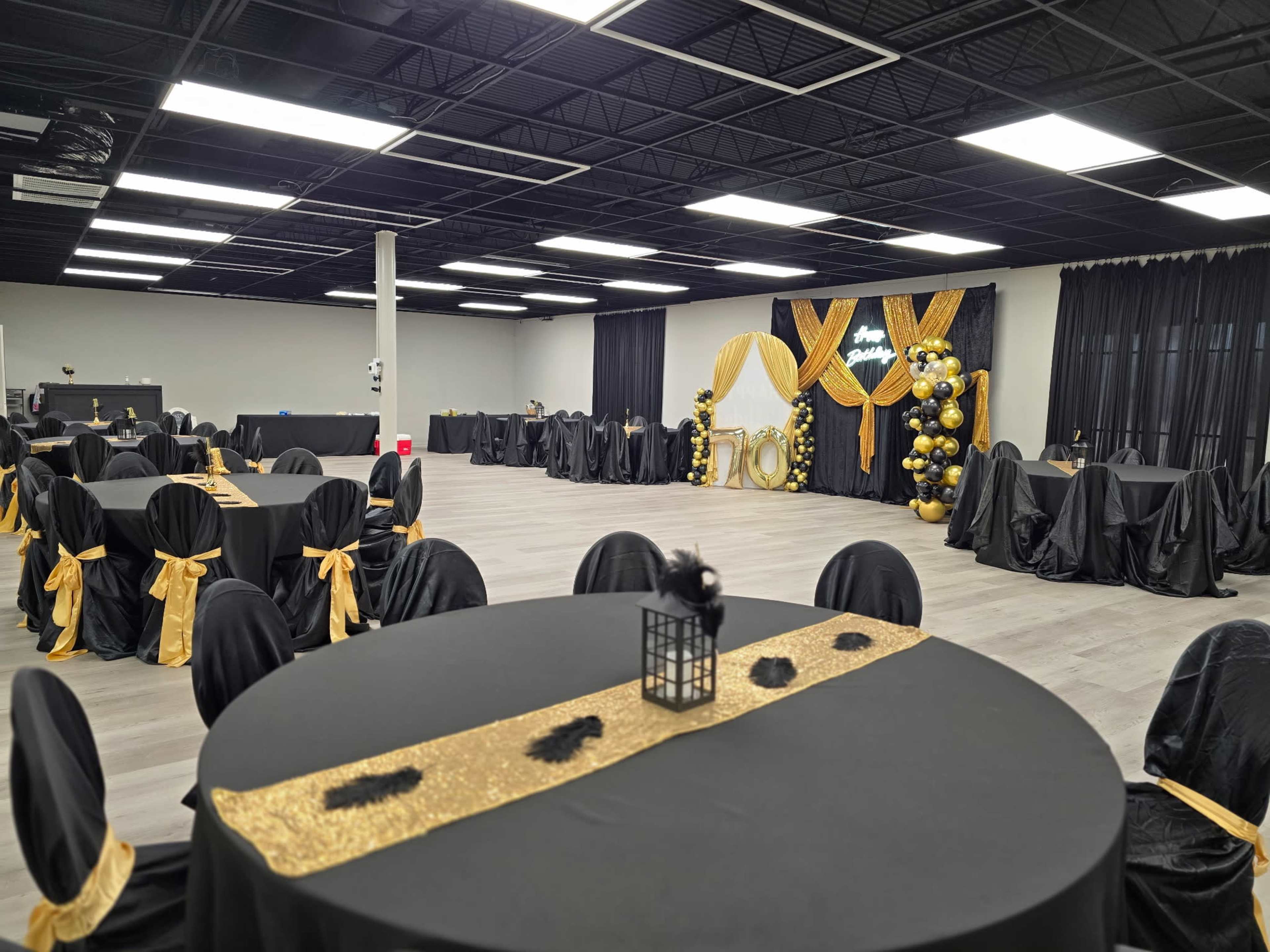 The image shows a spacious event hall set up with black and gold decorations, featuring round tables covered in black tablecloths and adorned with golden accents.