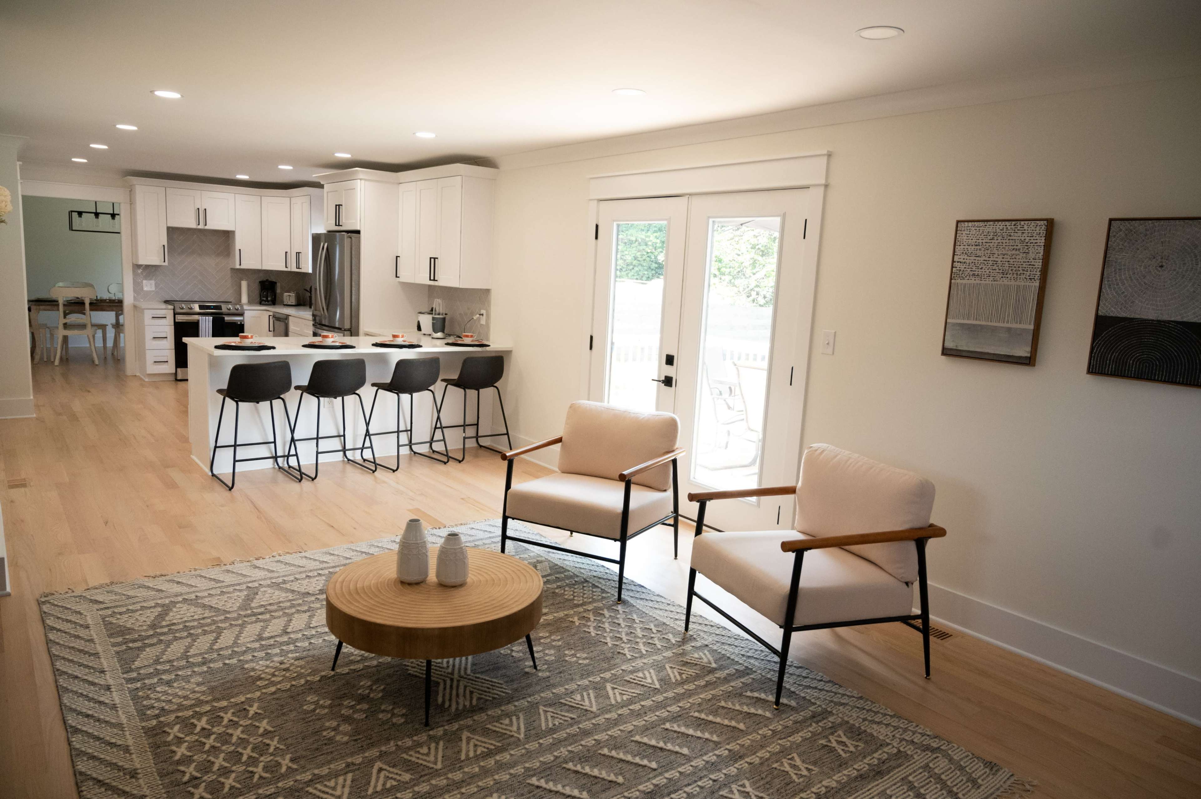 A modern living area features two armchairs and a circular coffee table with a geometric rug, adjacent to a kitchen with bar stools and white cabinetry.