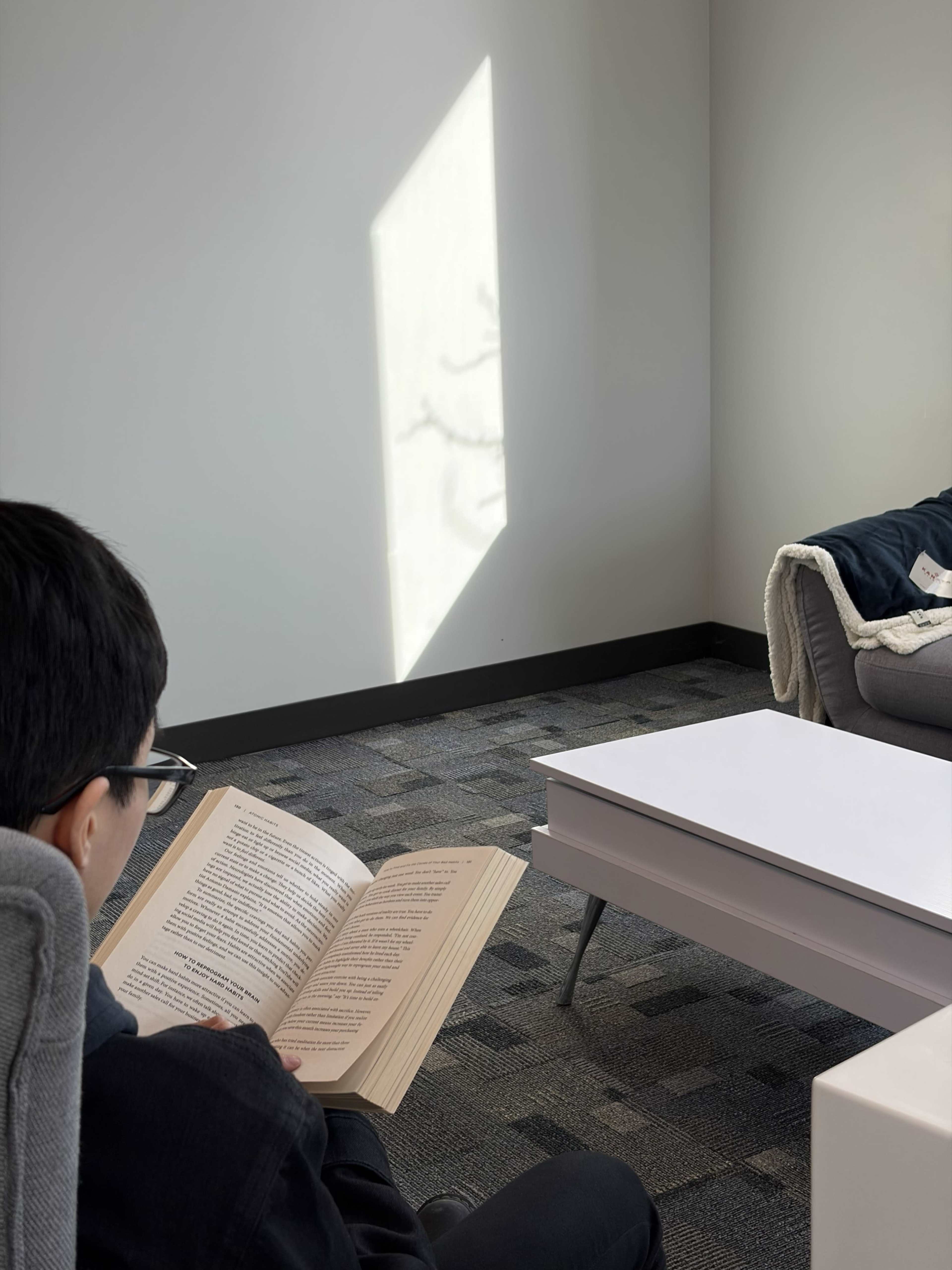A person reads a book while sitting on a chair in a modern, minimally furnished room with sunlight casting a shadow on the wall.