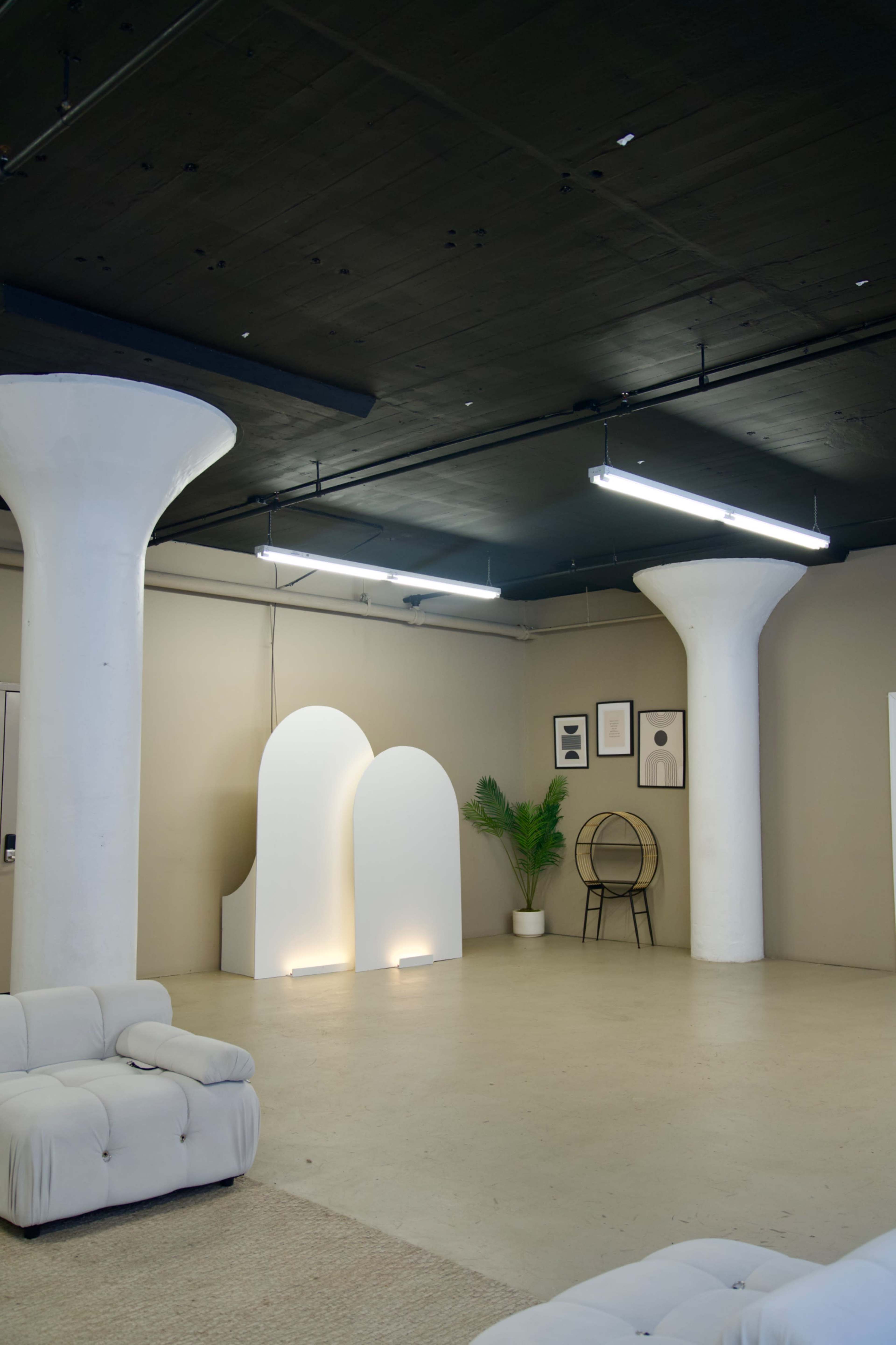 An open interior space features white pillars, minimalist arch-shaped decor, a potted plant, and modern furniture against plain walls.