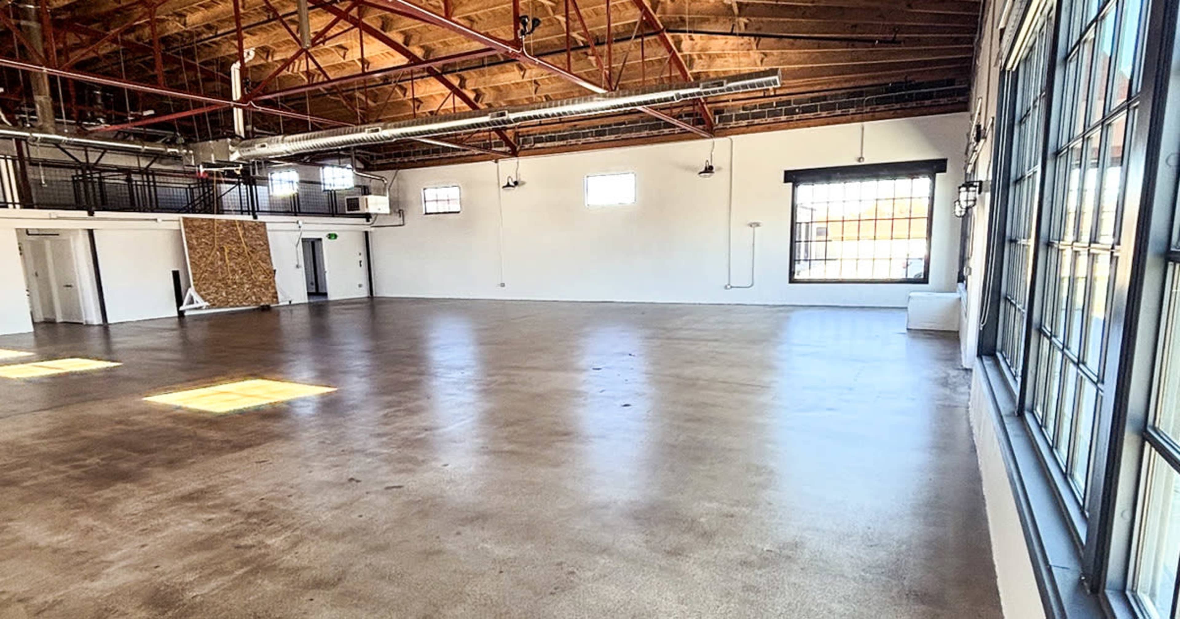 The image shows a spacious, empty room with a concrete floor, large windows, and exposed wooden beams in the ceiling.