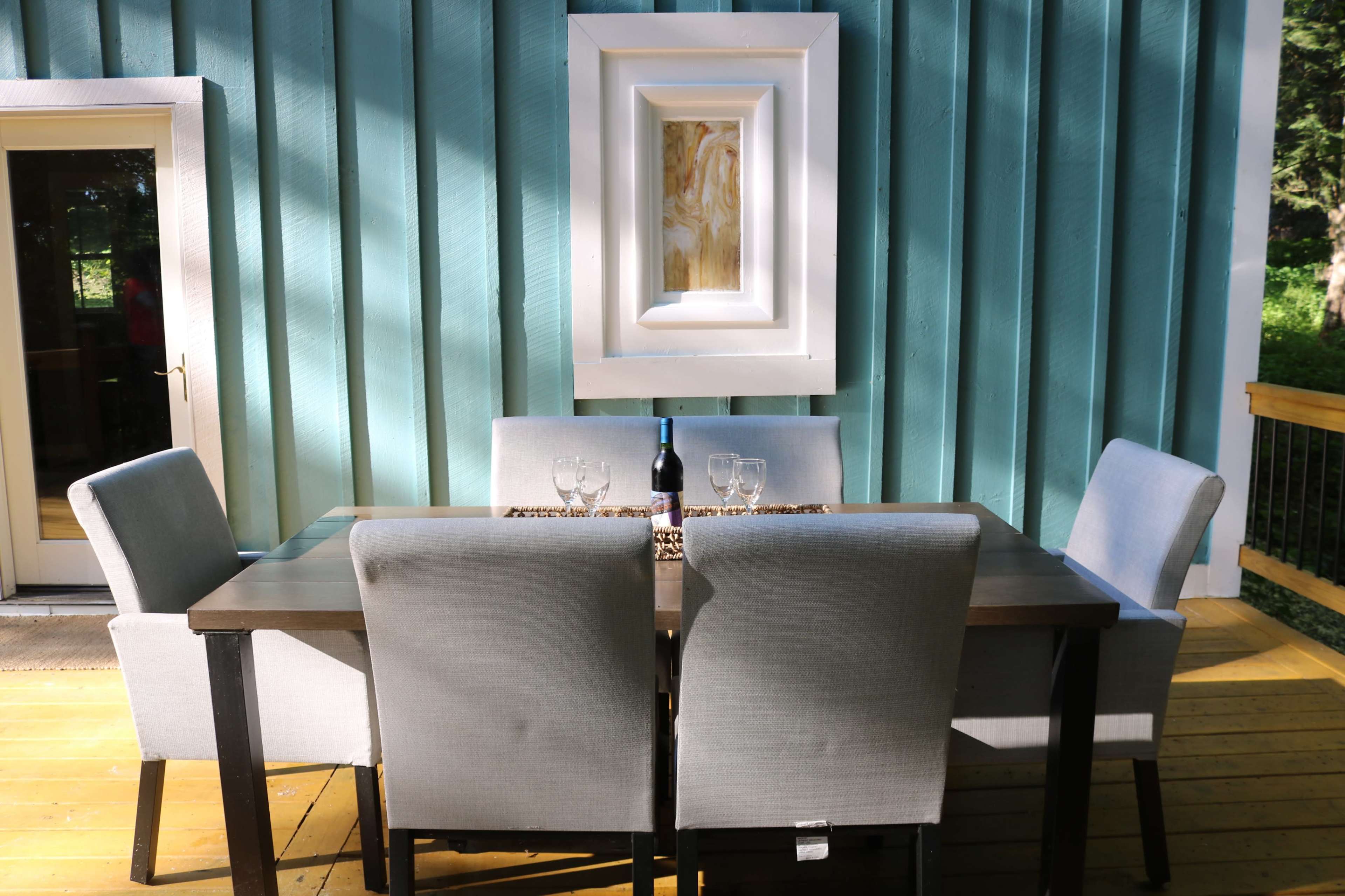 A dining table with four gray chairs is situated against a turquoise wall, featuring a framed artwork above and a bottle of wine with two glasses on the table.