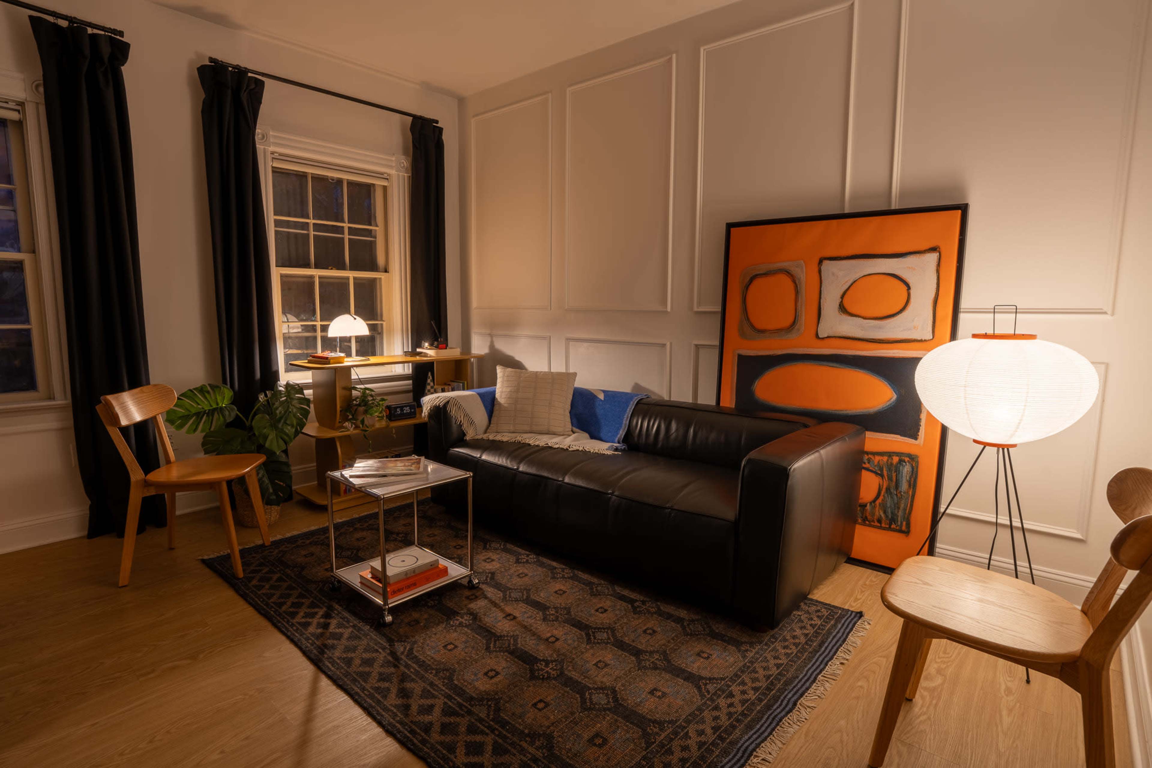 The image shows a cozy living room featuring a black leather sofa, a patterned area rug, a wooden side table, and a large piece of abstract art on the wall.