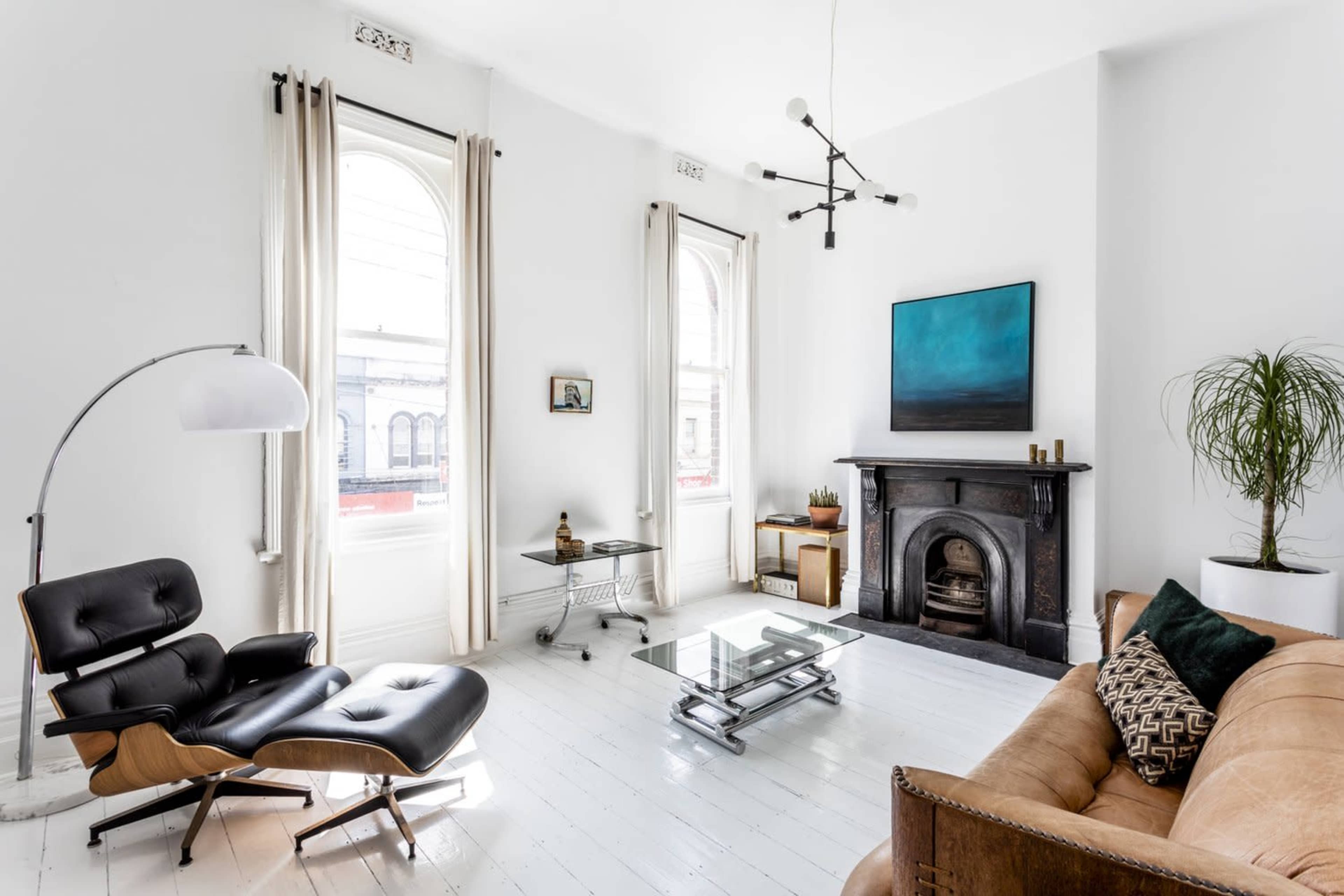 A modern living room features a leather sofa, an Eames chair, a glass coffee table, and a fireplace, with large windows allowing natural light to fill the space.